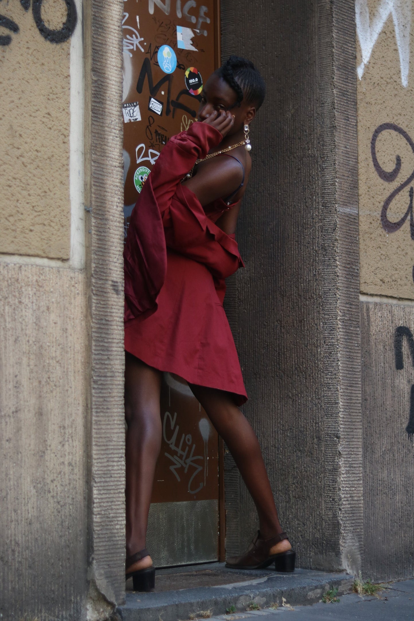 Urban Fashion Photography: Woman in Stylish Red Dress Posing by Graffiti-Covered Door – Trendy, Edgy, Street Style, Black Heeled Sandals, Bold Accessories, Vintage Dress, Cityscape Background.