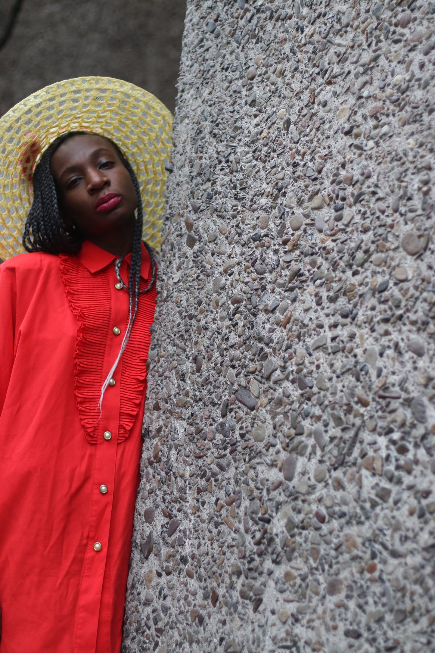 Fashionable model of African heritage wearing a vintage vibrant red button-up shirt with ruffle detailing, complemented by a straw hat, leaning against a grey wall