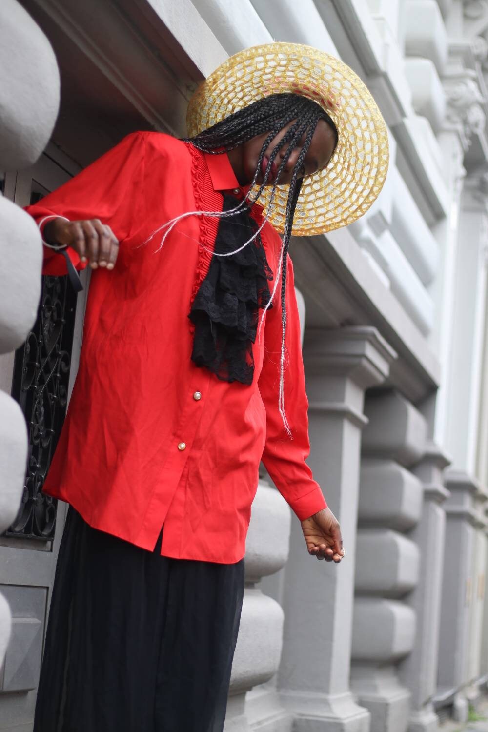 Fashionable person wearing a red blouse and straw hat with long braids stands against an urban architectural backdrop.