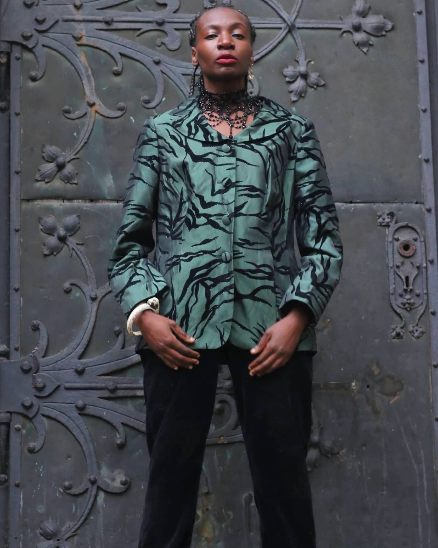 Model of African heritage wearing a vintage green and black zebra-print blazer, standing confidently in front of an ornate dark green wrought-iron door with floral motifs.