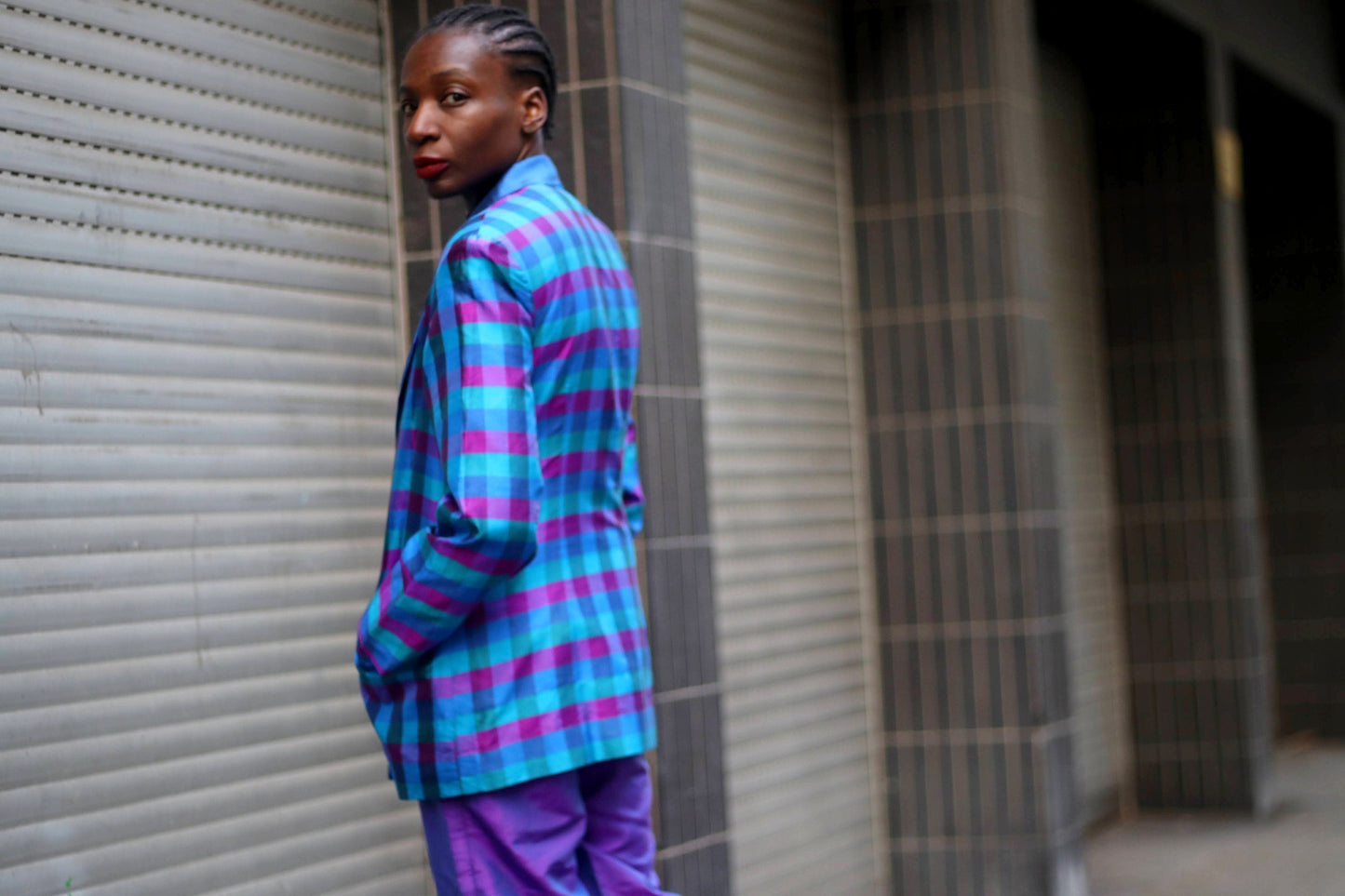 A stylish woman in a vintage checkered blue and purple blazer paired with purple pants poses confidently in an urban setting. Both hands are in the blazer pockets. The fashion-forward outfit stands out against the industrial background