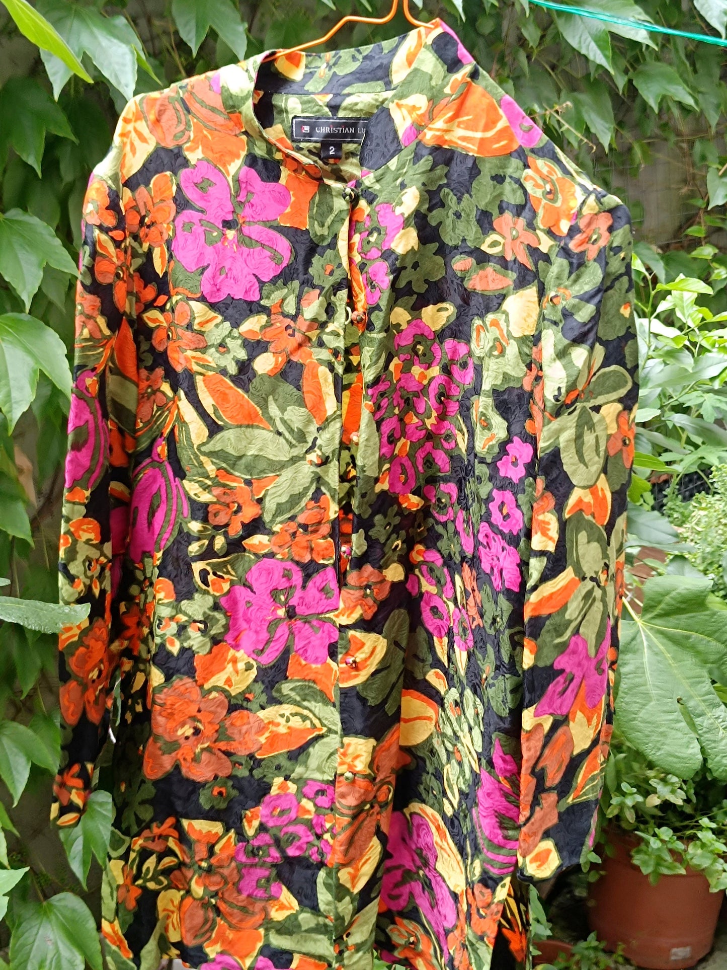 The Christian Lu silk blouse, inspired by the qipao, is displayed on a hanger, highlighting its traditional mandarin collar, statement floral print, and lightweight silk drape.