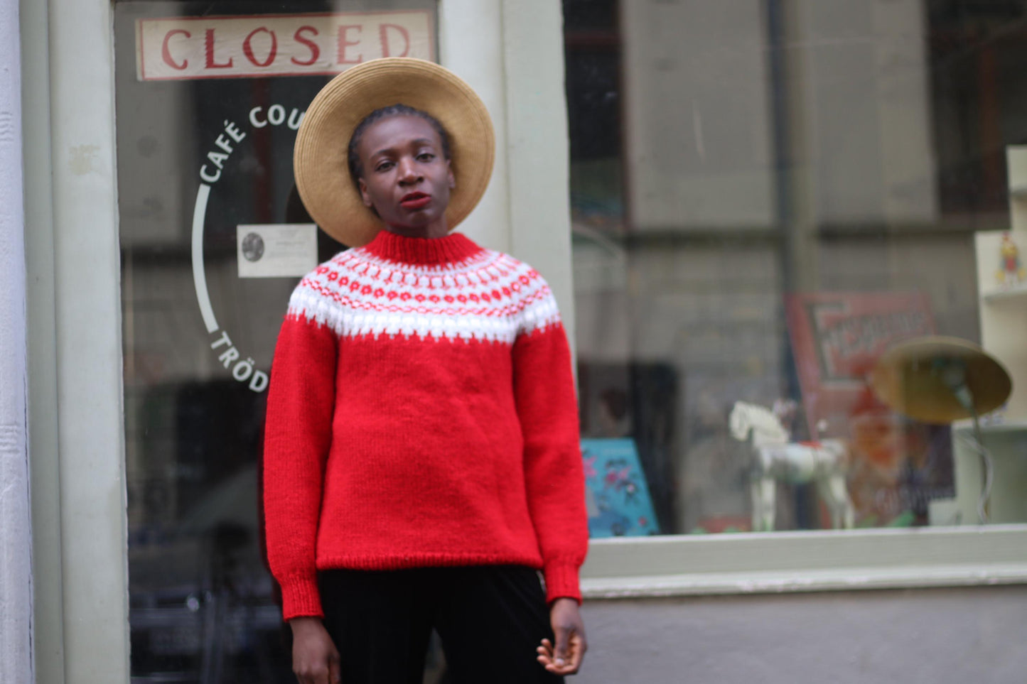 Black female model in a vintage Scandinavian red sweater with white patterns and wide-brimmed hat in front of a shop window with a closed sign. The chunky handmade sweater has a high neck silhouette and long sleeves