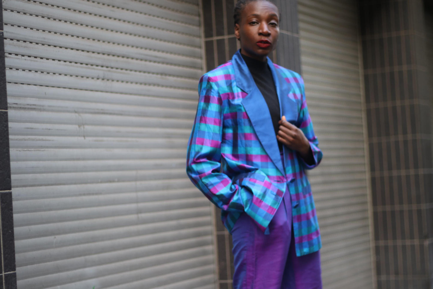 A stylish woman poses confidently in an urban setting, wearing a vibrant checkered blue and purple vintage blazer paired with purple pants. The fashion-forward outfit stands out against the industrial background, offering a striking contrast.