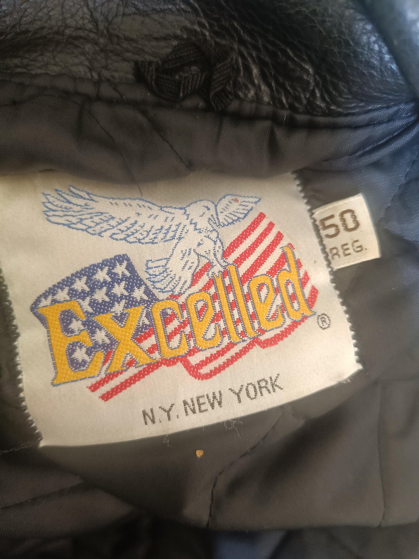 Close up of the brand tag of a vintage black Leather Jacket by the brand Excelled. The tag reads : Excelled N.Y New York and also includes size information: 50 Reg T