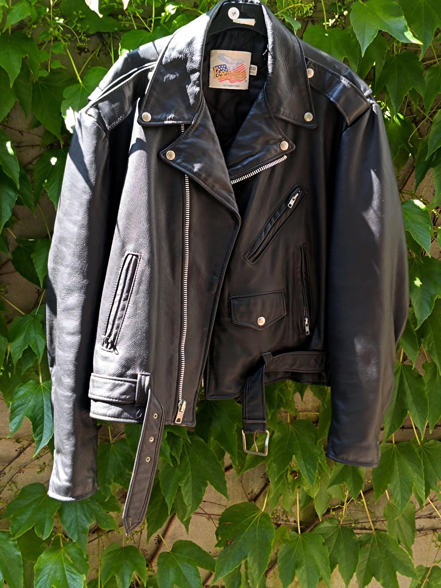 A vintage black Leather Jacket by the brand Excelled photographed hanging and against a green leafy background. The trendy biker jacket is from the 90s