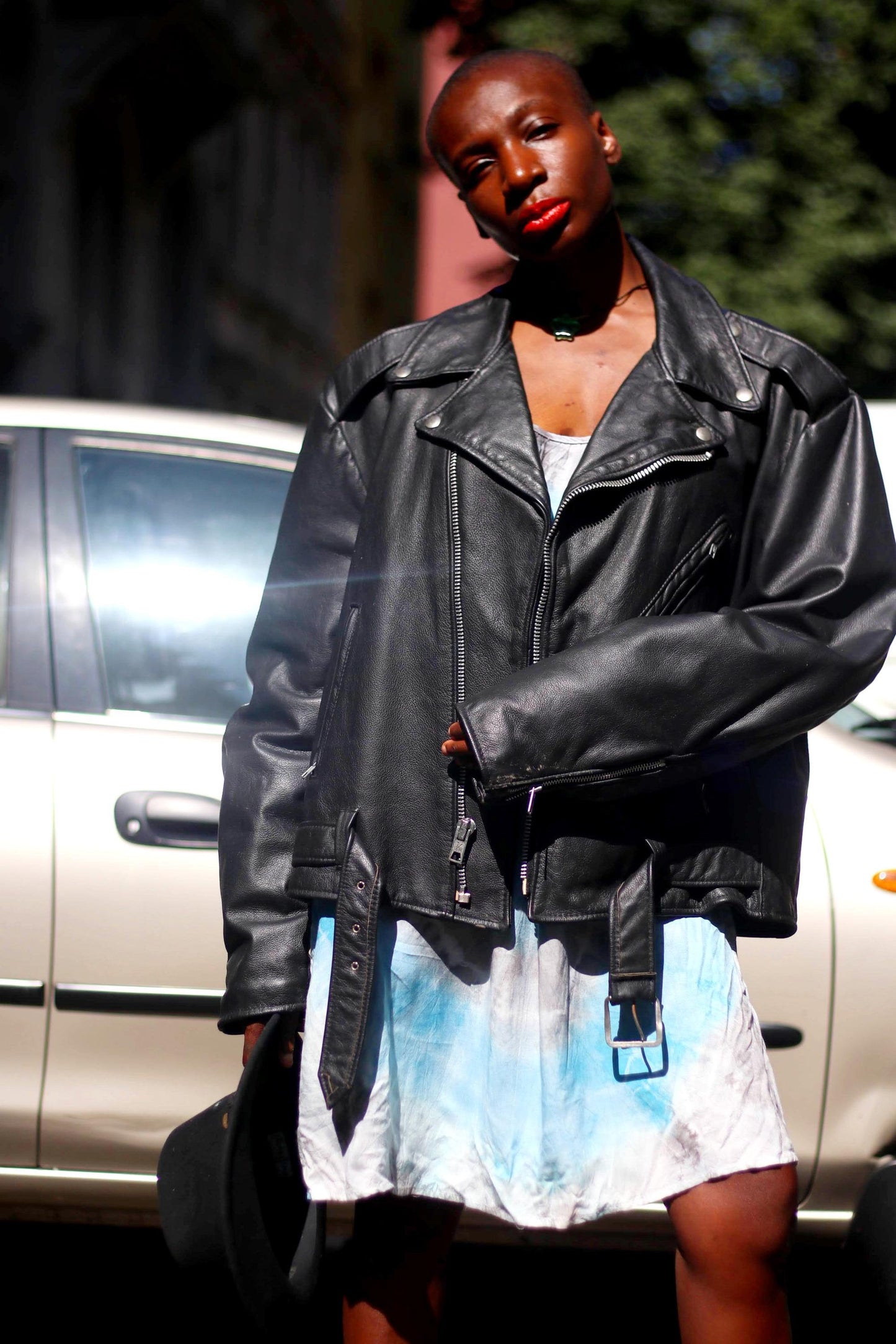 A female model of African heritage wearing an oversized black biker jacket from the 90s over a trendy blue tie dye spaghetti-strapped dress photographed in front of a silver car in an urban setting