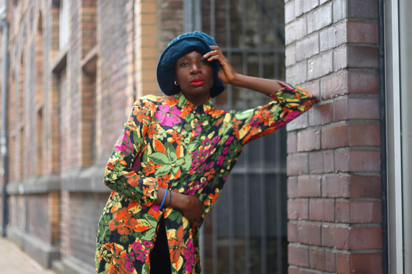 A stylish model leans against a brick wall wearing a vintage silk blouse inspired by traditional cheongsam designs. The floral print pops against the rich black background. she also wears a blue hat