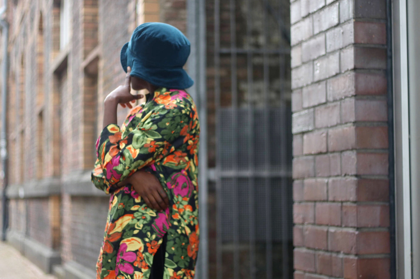 A model poses in a vintage Christian Lu silk blouse with a mandarin collar and bold floral print in pink, orange, and green is photographed near a brick wall. The draped fit and long sleeves create a striking, retro-chic silhouette.