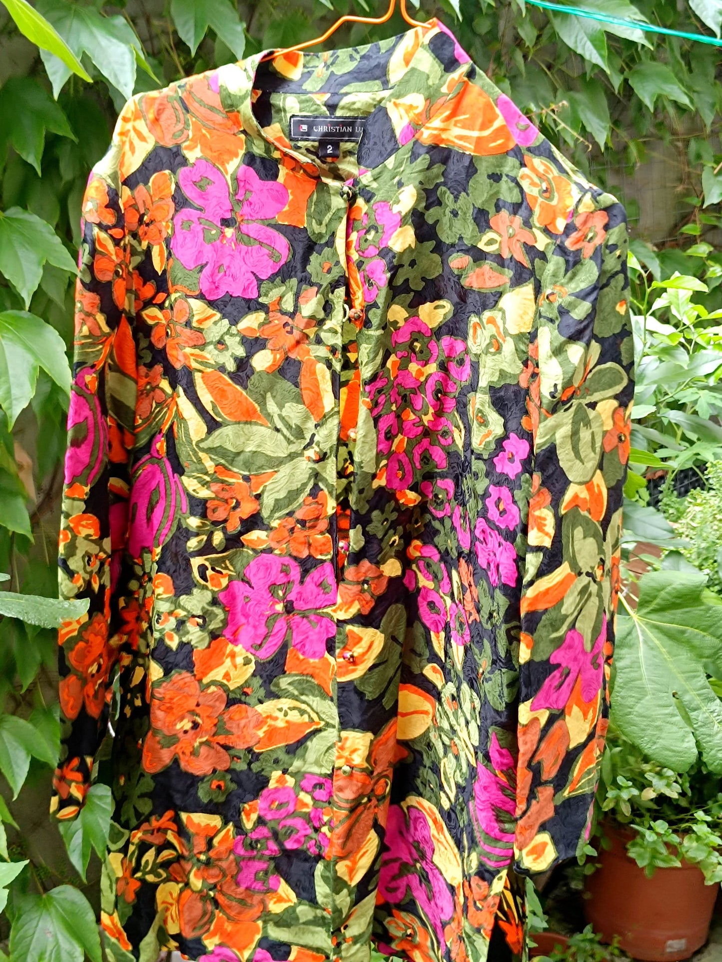 A vintage Christian Lu silk blouse with a mandarin collar and delicate buttons hangs against lush greenery, showcasing its vivid floral print in pinks, oranges, and greens.
