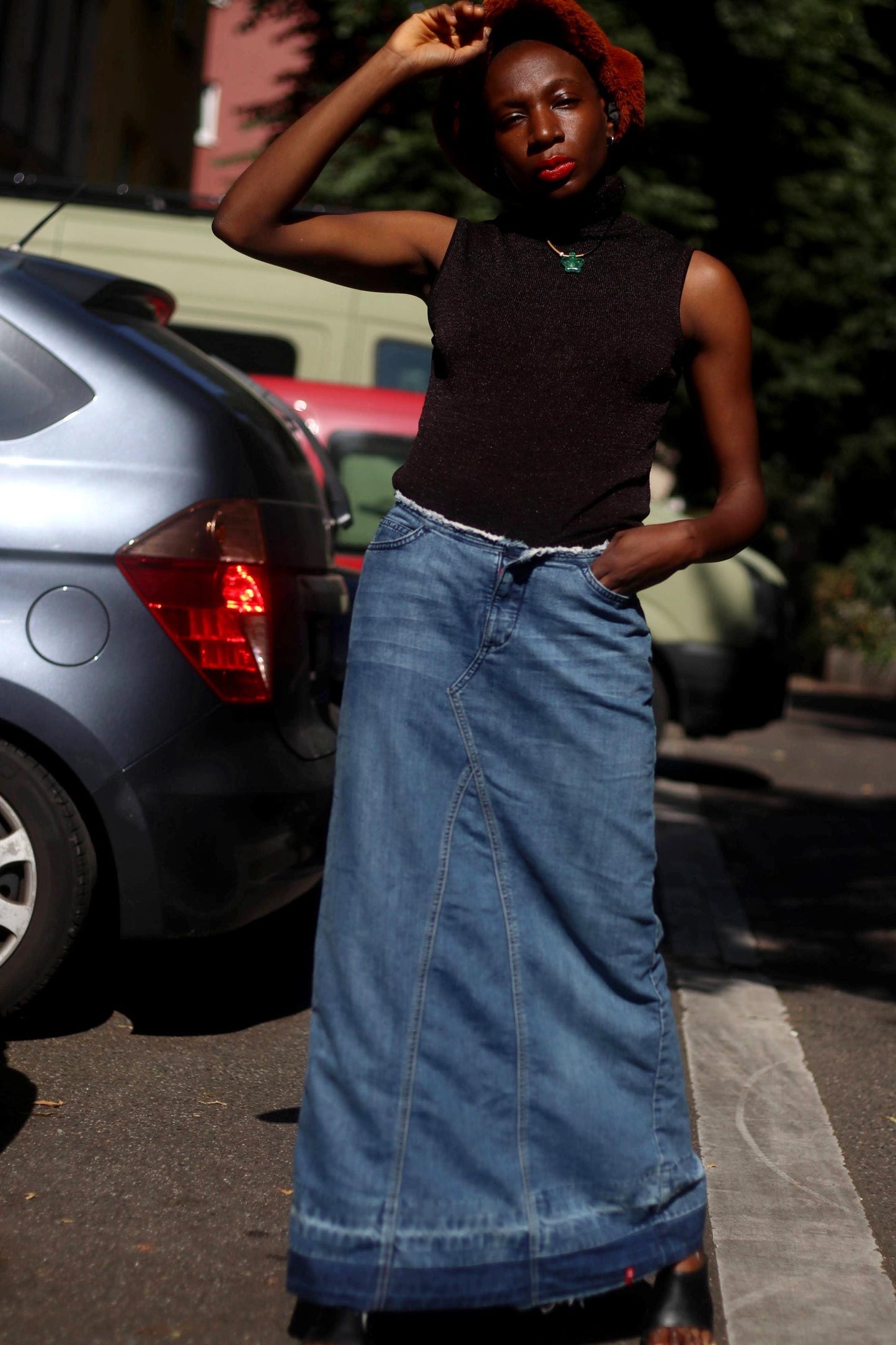 A female model in an Esprit Maxi Skirt with black sleeveless turtleneck and an oversized fuzzy brown newspaper boy hat is photographed outdoors in the street with cars in the background
