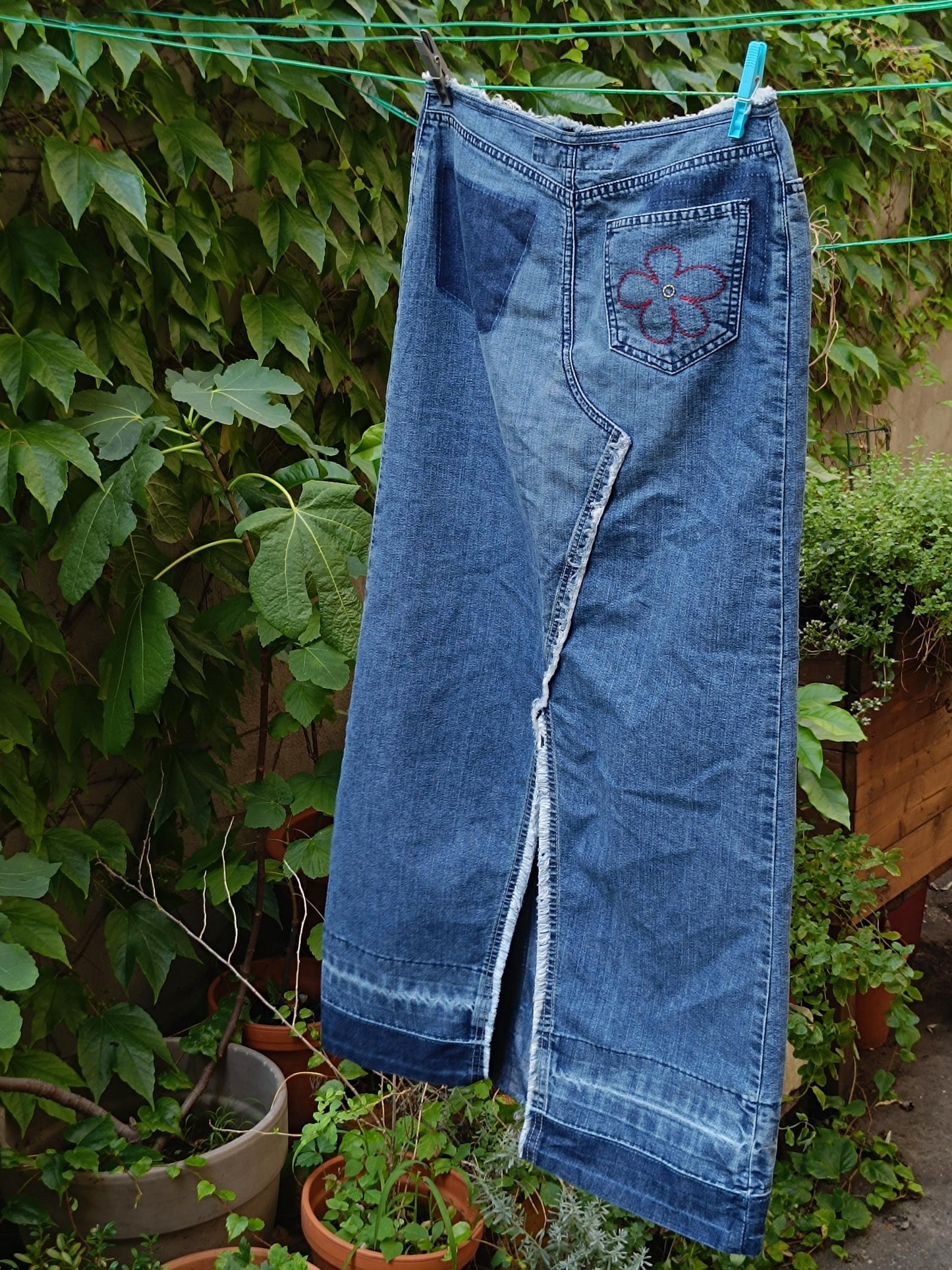 The back view of A y2k Esprit Maxi Skirt is hanging from a clothing line against a background of green plants. The image shows the floral embroidered details of the Vintage Low-Waisted Hippy Blue Jeans Skirt .