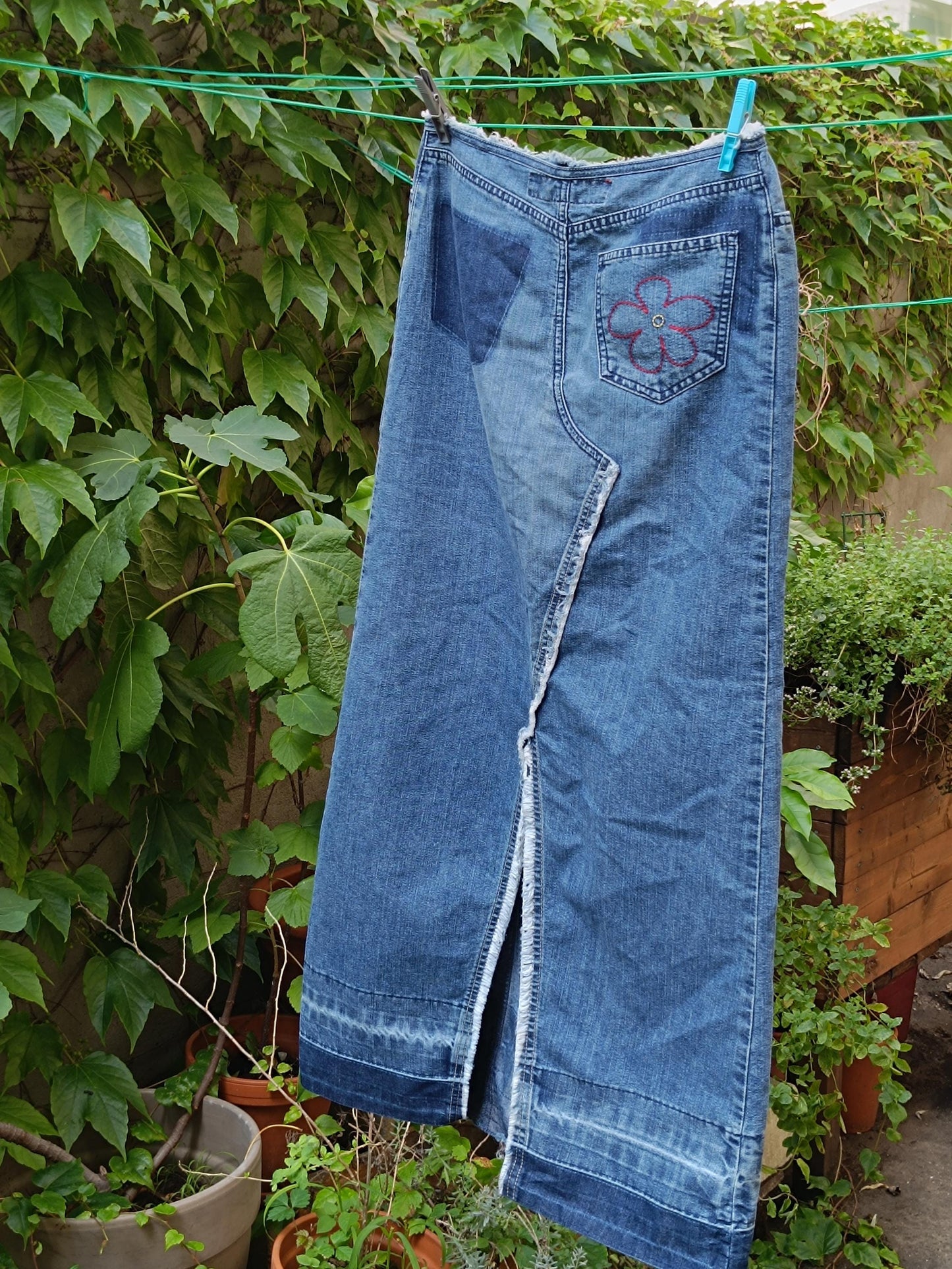 A y2k Esprit Maxi Skirt is photographed hanging from a clothing line against a background of green plants. The image shows the patchwork details of the Vintage Low-Waisted Hippy Blue Jeans Skirt .