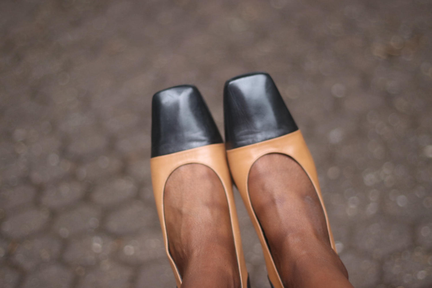 A person wearing two-toned brown and black flats photographed against a cobblestone surface.
