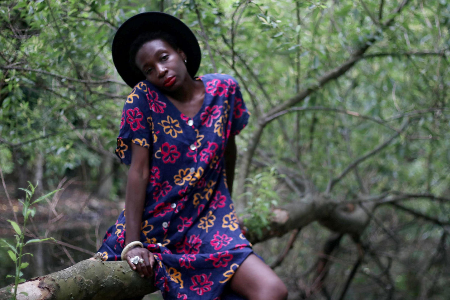 A black woman in vintage blue floral dress sitting on a tree branch. She is wearing a black hat.