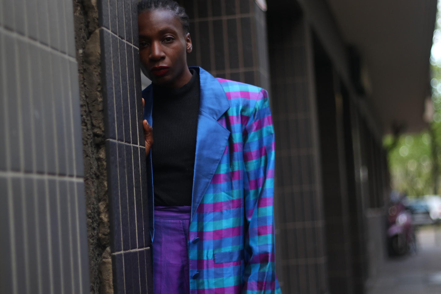 A stylish woman poses confidently in an urban setting, wearing a vibrant checkered blue and purple vintage blazer paired with purple pants. She wears a black turtle neck under the blazer. The fashion-forward outfit is trendy for work or play