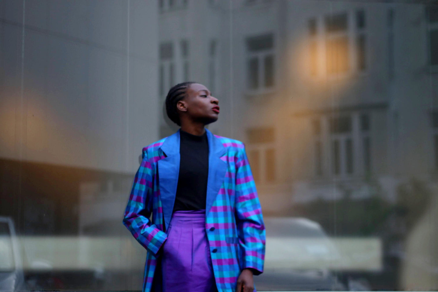 A black woman is photographed leaning against large glass window with an arty reflection of buildings . She wears a vintage checkered blue and purple blazer with purple pants. Her hands are in her pockets. The fashion-forward outfit is all vintage