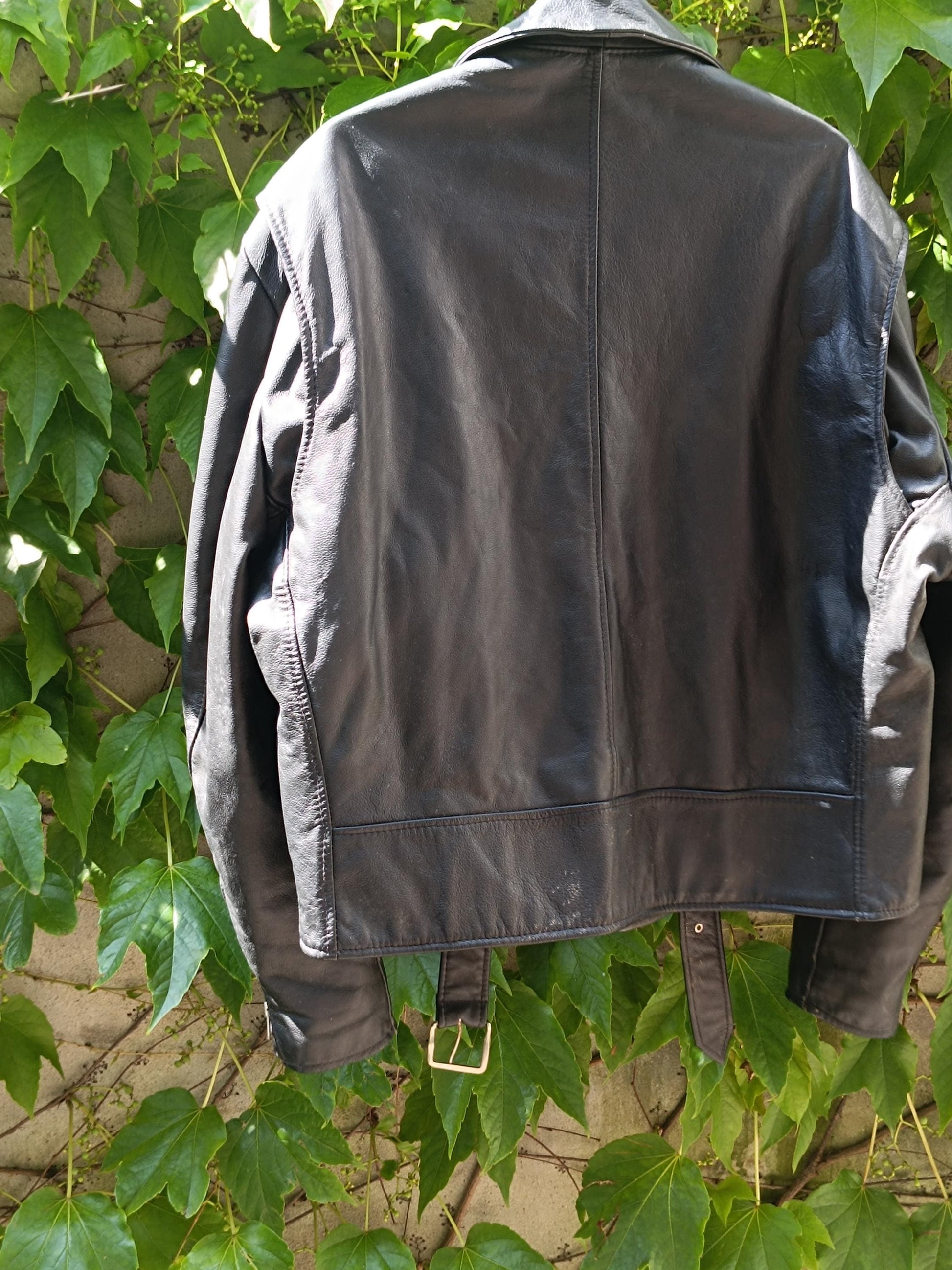 The back of a vintage black Leather Jacket by the brand Excelled is photographed hanging and against a green leafy background. The trendy biker jacket is from the 90s