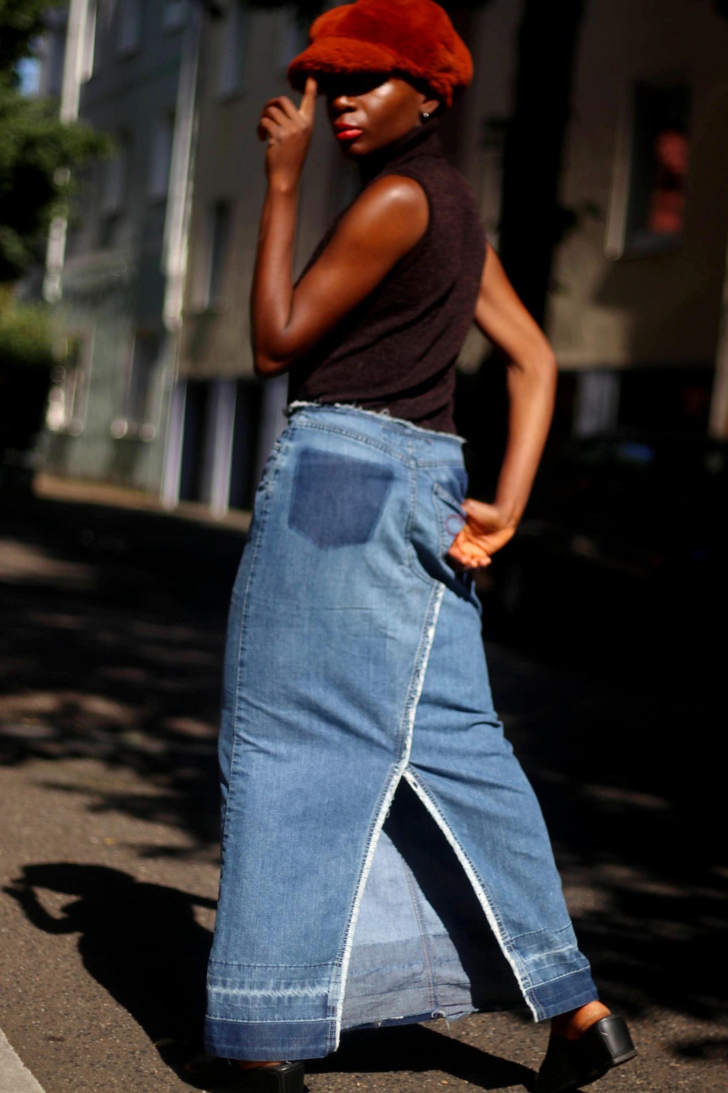 The back view of female model in an Esprit Maxi Skirt with black sleeveless turtleneck and an oversized fuzzy brown newspaper boy hat outdoors in the street with cars in the background. the image showcases the patchwork details and the floral embroidered element