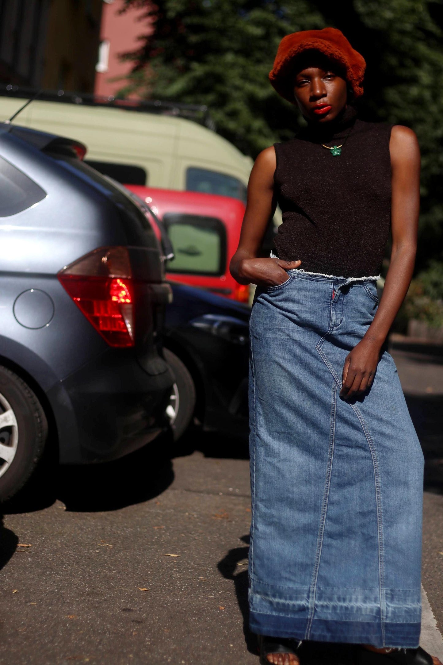 A female model in an Esprit Maxi Skirt with black sleeveless turtleneck and an oversized fuzzy brown newspaper boy hat is photographed outdoors in the street with cars in the background