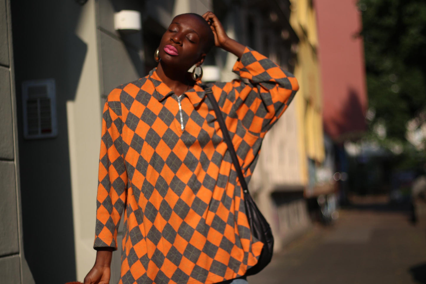 Black female model in a vibrant vintage orange and grey checkered sweater paired with denim shorts outdoors and a black hobo bag. Her eyes are closed and one hand is on her head Expert in street fashion, casual outfit inspiration, and urban style.