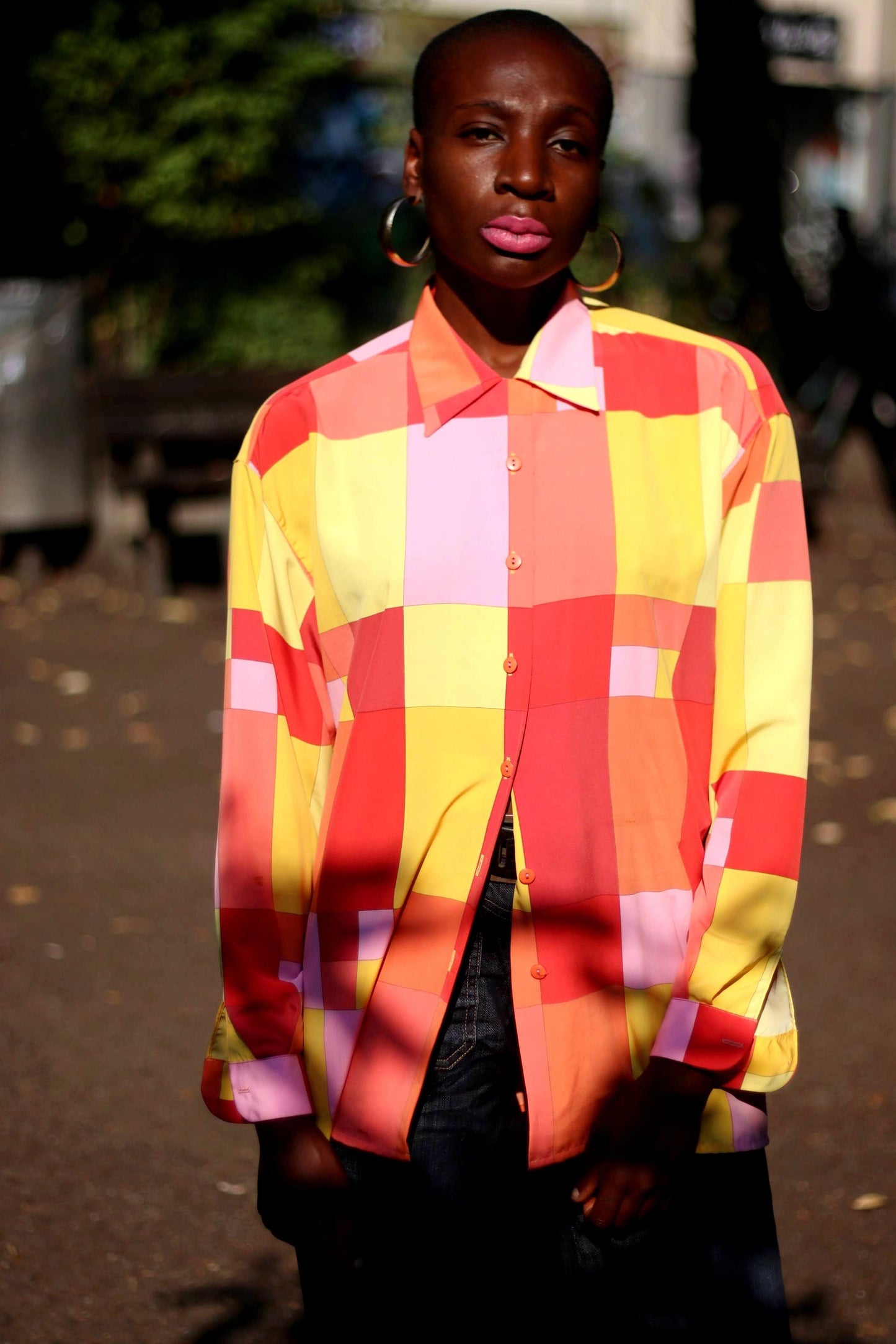 Model in a vibrant, vintage colorful checkered shirt with shades of red, pink, yellow, and orange, posing outside against a green background near some benches concept in outdoor setting.
