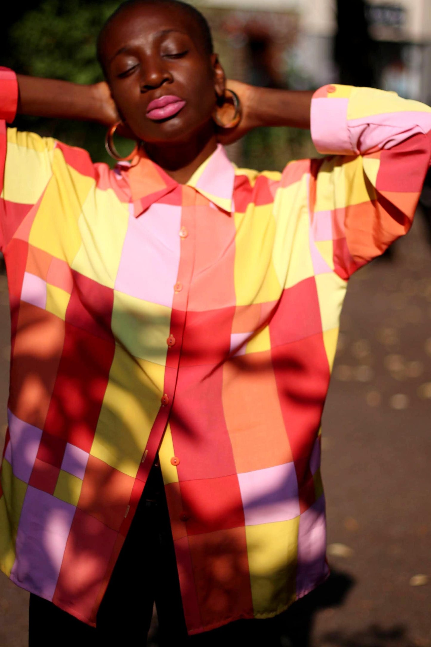 A female model of African heritage in a colorful checkered shirt with shades of red, pink, yellow, and orange, posing with eyes closed and arms raised behind head. The blouse is from the 80s and vintage. Fashion & style concept in outdoor setting.