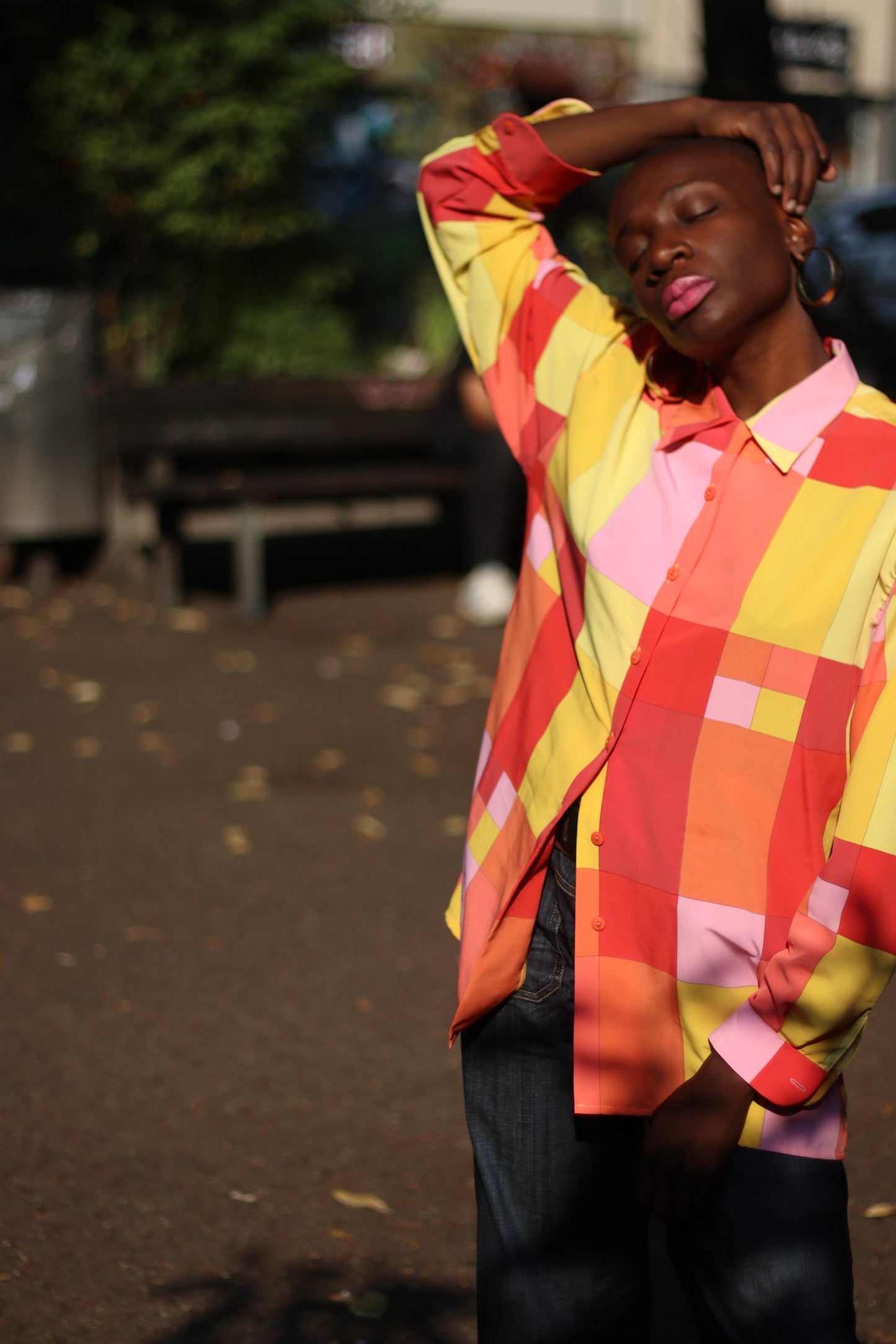 A female model of African heritage in a colorful checkered shirt with shades of red, pink, yellow, and orange, posing with her eyes closed and one hand on the head. The vintage 80s blouse by the brand Romazzino is paired with high waisted jeans