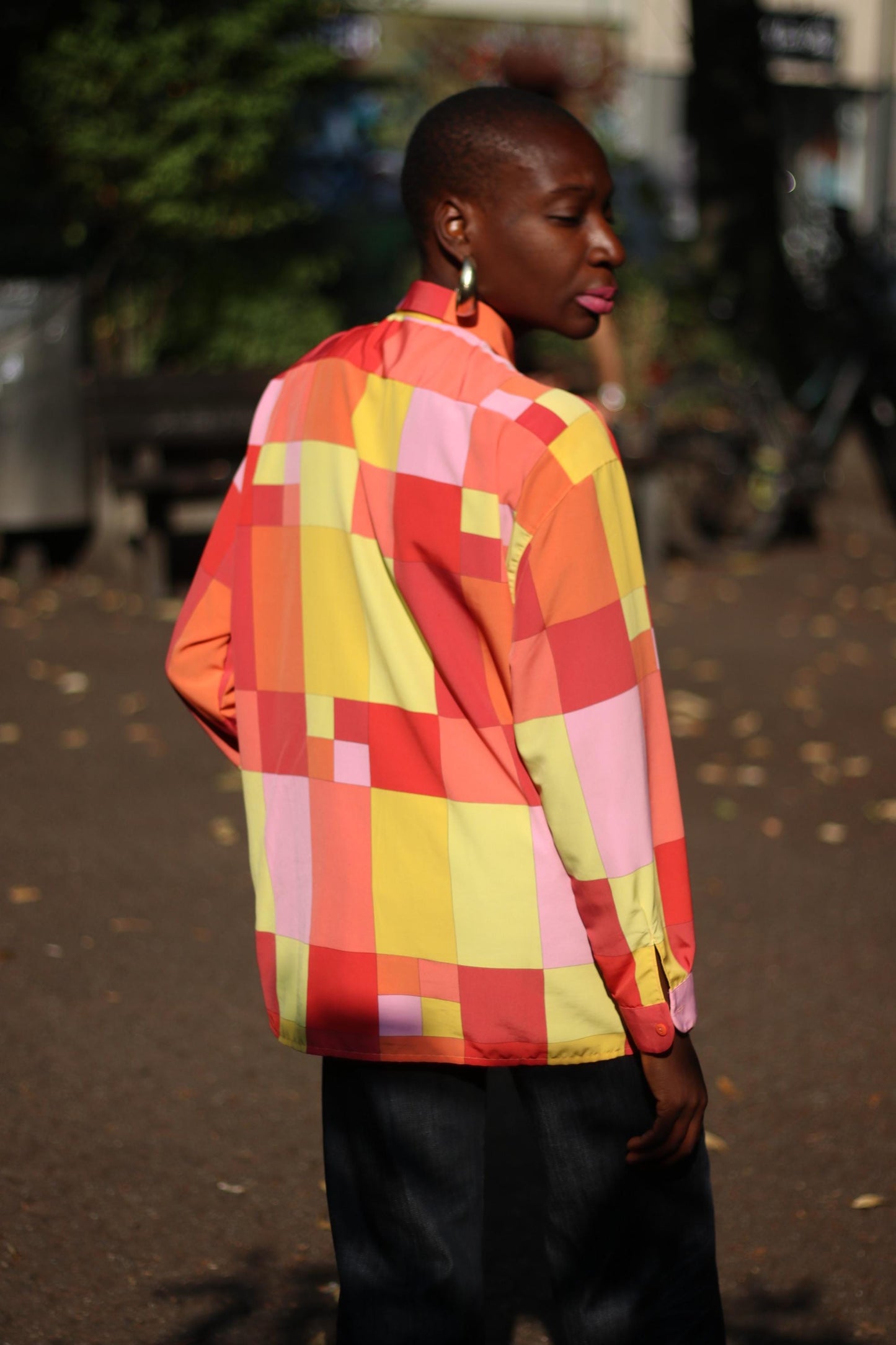 A female model of African heritage in a colorful checkered shirt with shades of red, pink, yellow, and orange, posing with her back to the camera. The vintage blouse is from the 80s and by the brand Romazzino Paris.