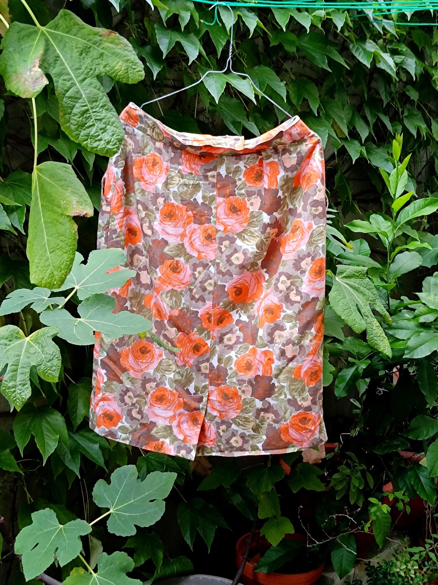 An 80s Orange Floral Skirt photographed hanging from a hanger with green plants in the background. The image showcases the bold print on the vontage knee-length Skirt