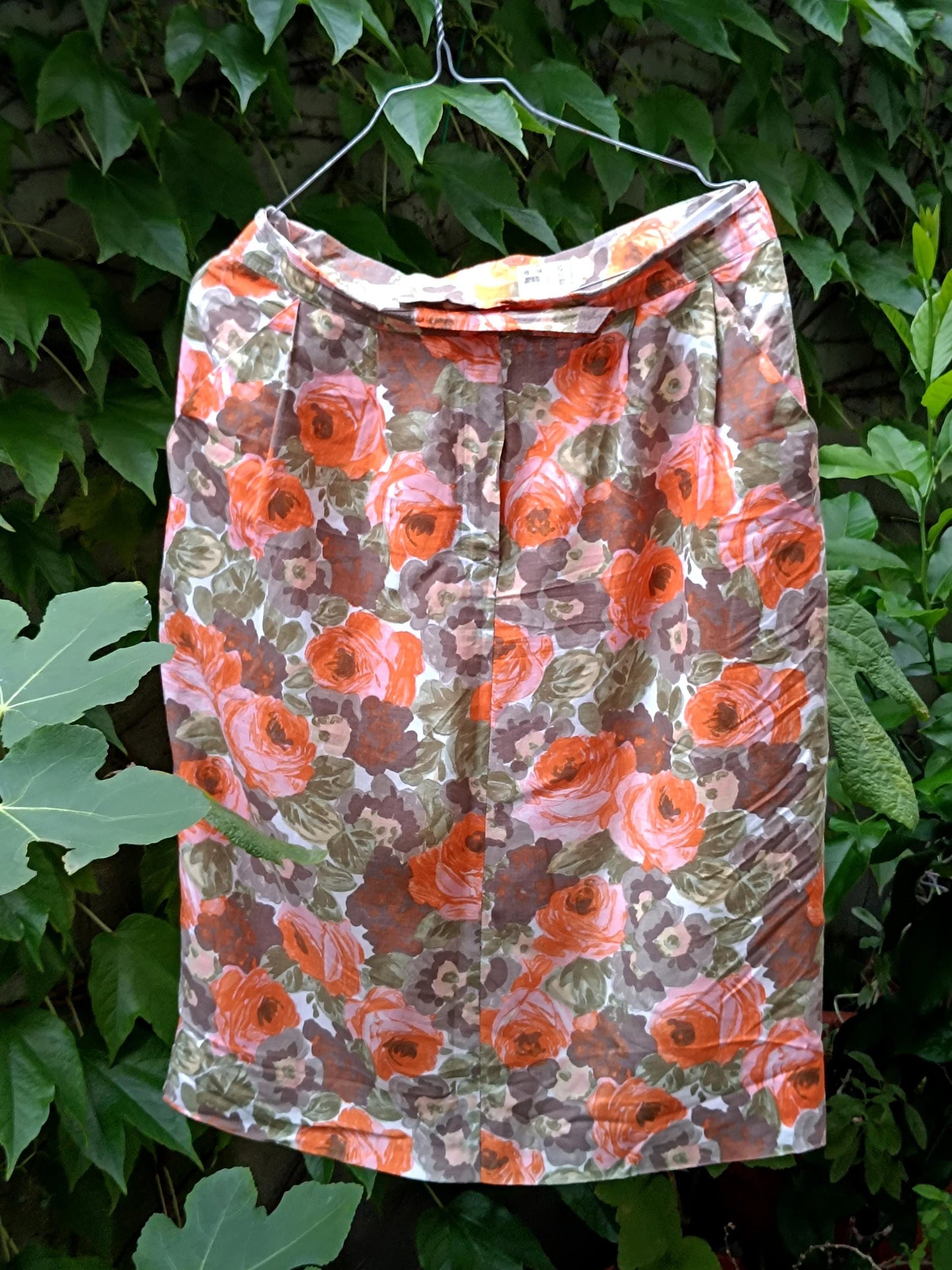 An artistic showcase of a vintage floral from the 80s shot outdoors against a leafy background to highlight the bold orange blooms on a retro knee-length skirt