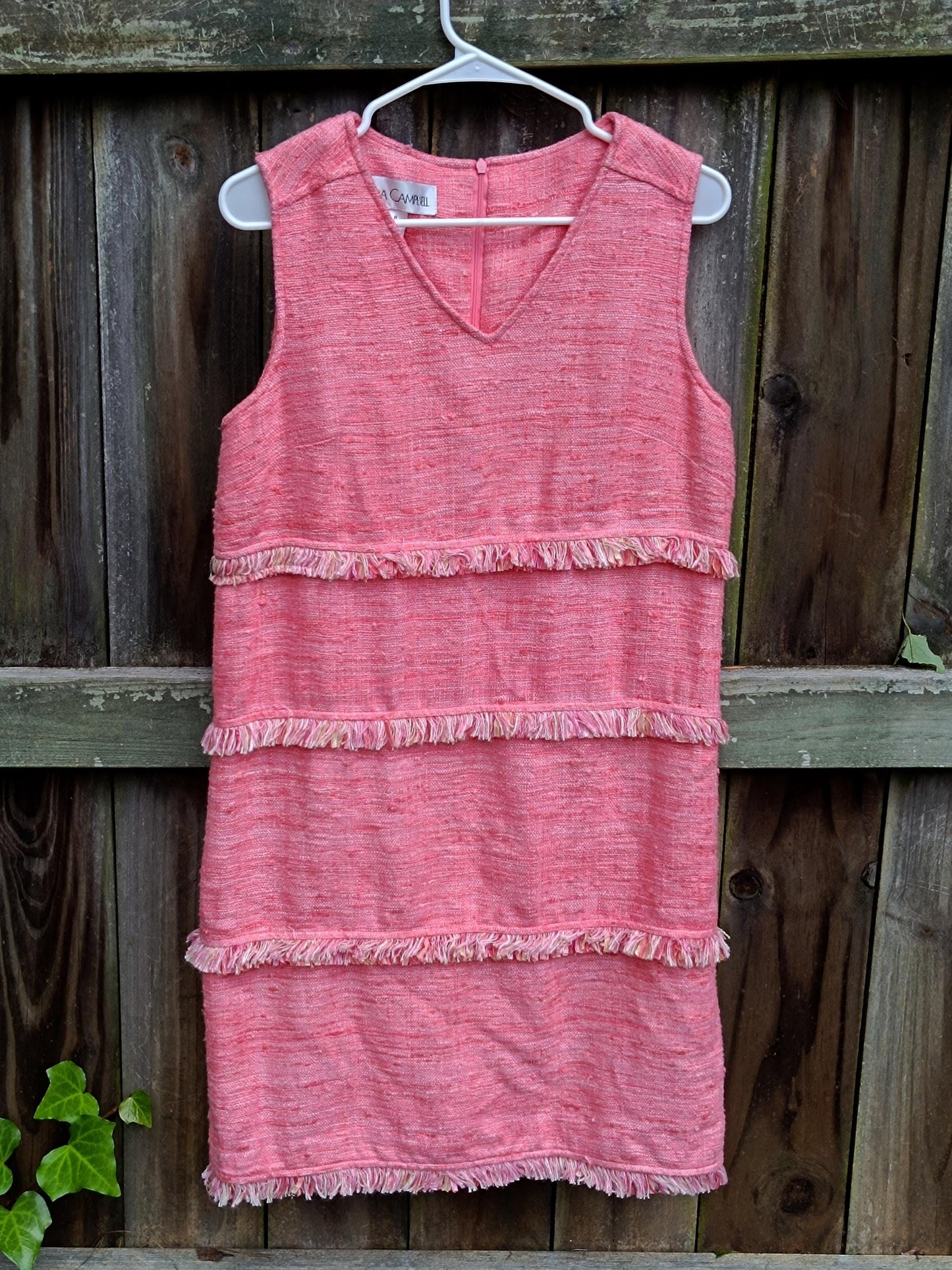 A pink sleeveless dress by Sara Campbell is photographed against a wooden fenced background with leafy details. The chic vintage dress with tiered fringed details was made in the 90s
