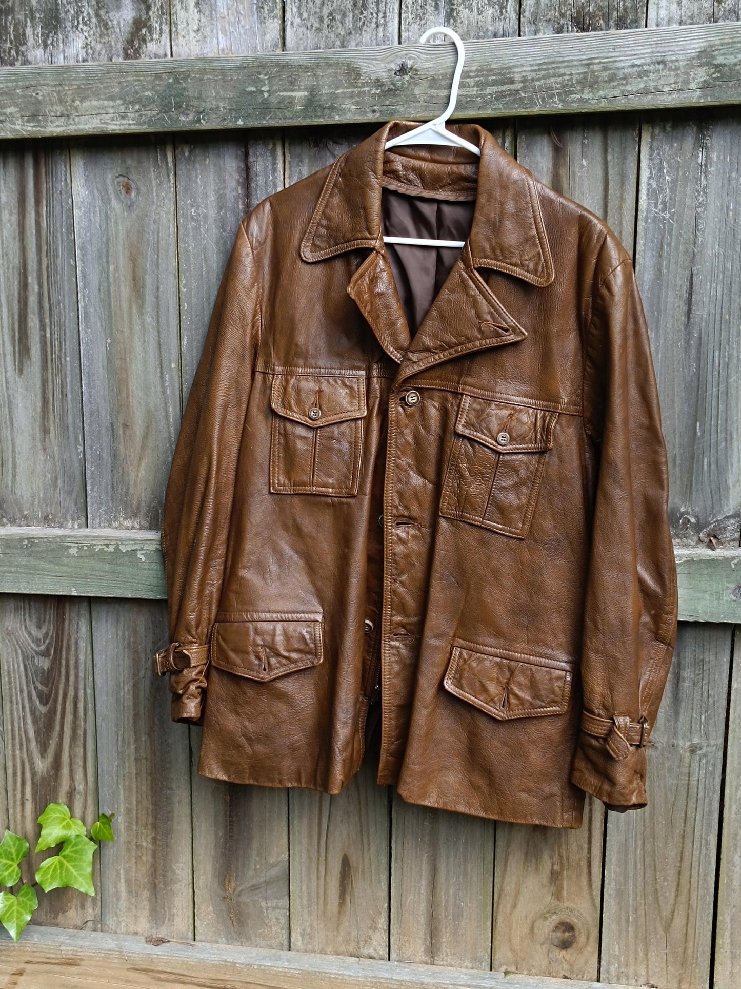 A 90s Vintage Leather Jacket in brown is photographed hanging on a hanger Infront of a brown rustic wooden fence. The image highlights the huge and minimalist cool silhouette