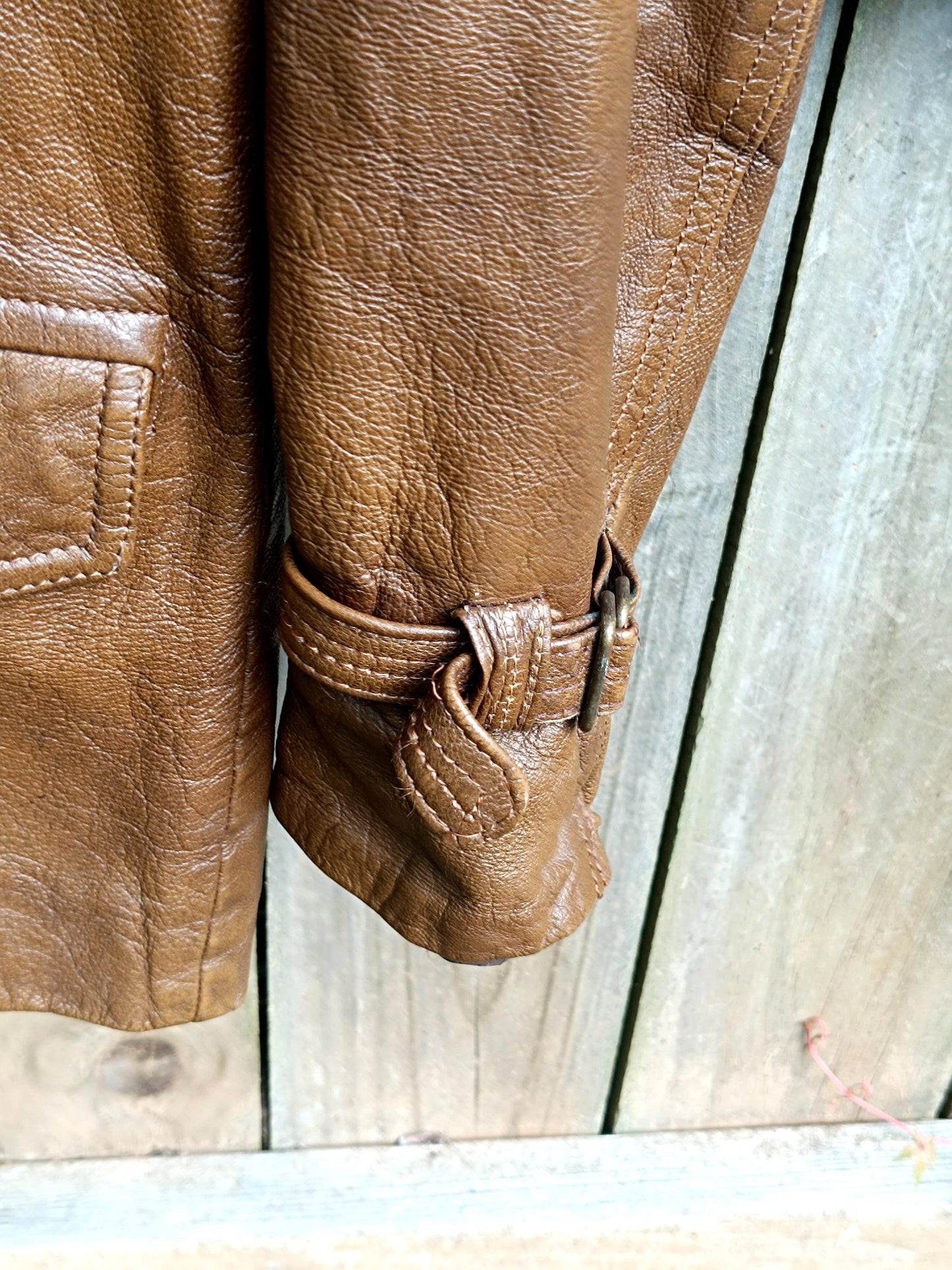 A detailed shot of the sleeves of a 90s Vintage Leather Jacket in brown photographed against a brown rustic wooden fence.The image highlights the hue and details of the sleeves