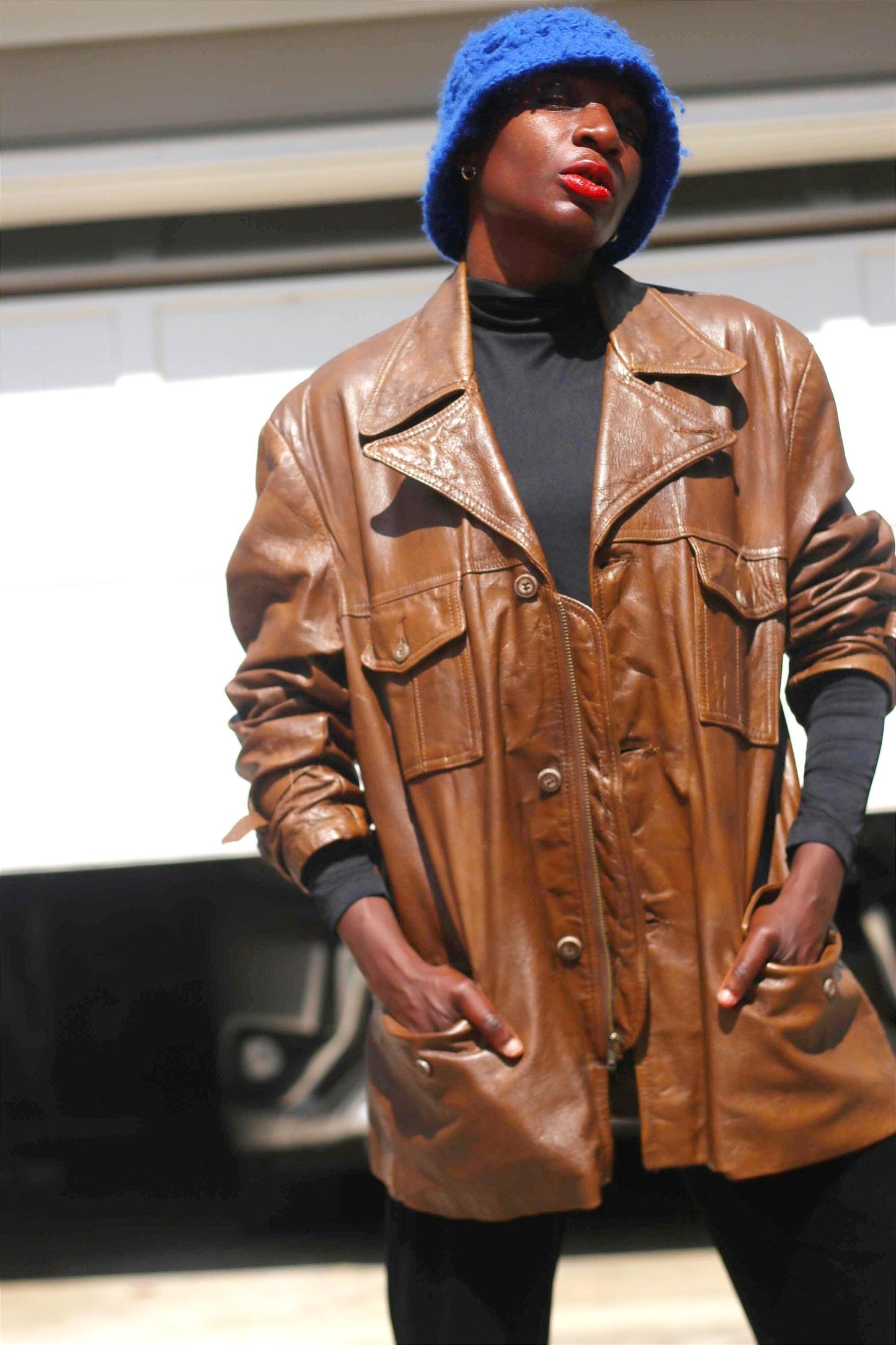 A women wearing a 90s Leather Jacket over a black turtleneck with a blue knitted bucket hat is photographed outdoors standing Infront of a white wall. Both hands are inside the Vintage Brown Unisex Leather Coat . The vibe is sleek Minimalist 90s streetwear