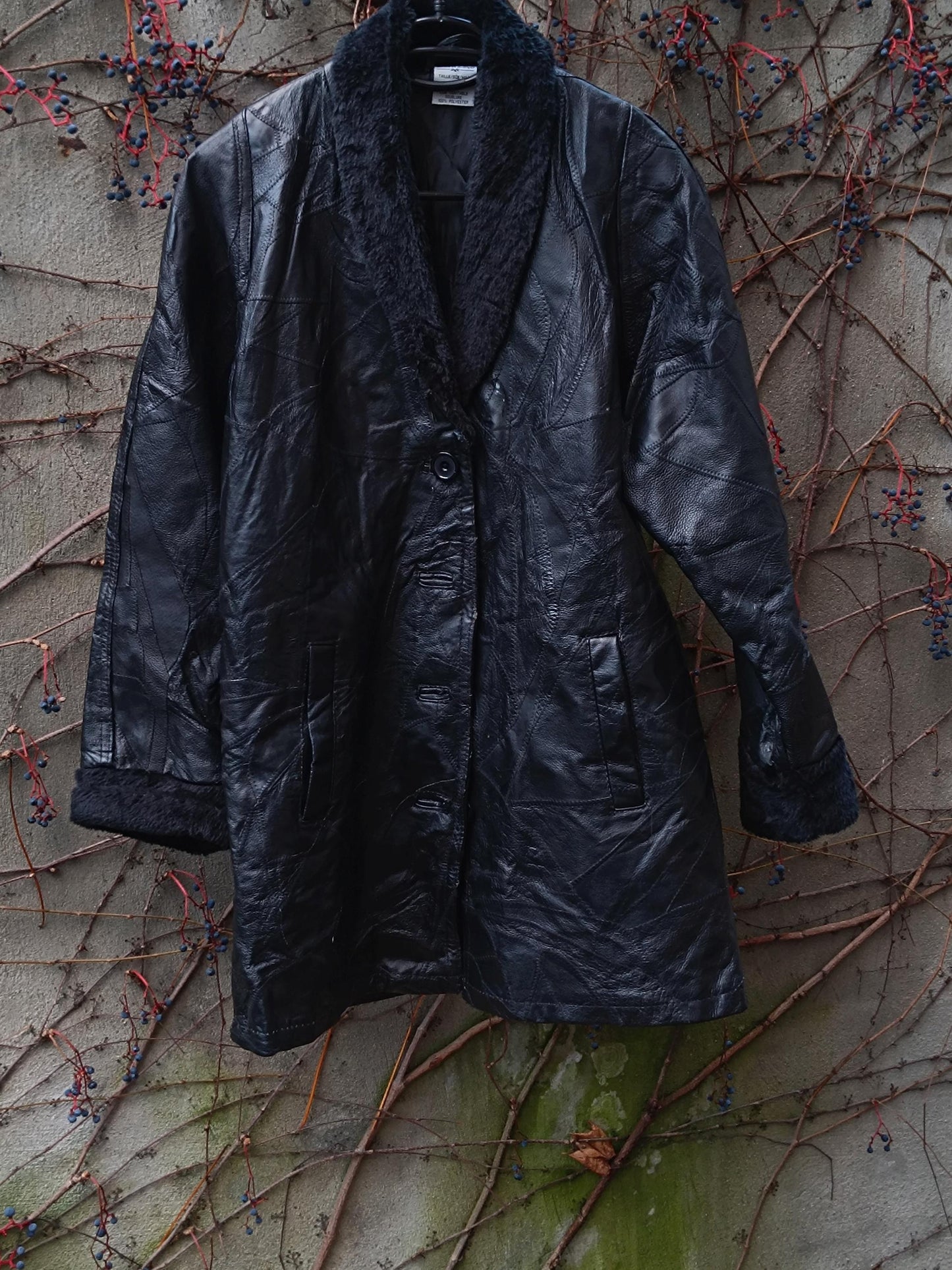 90s vintage black leather jacket with a faux fur trimmed collar and sleeves by the brand Anne De Lancay. The boho chic women's Coat is photographed hanging outdoors in a garden against a grey wall with browning twines and leaves. The patchwork design gives off a rock and roll boho vibe