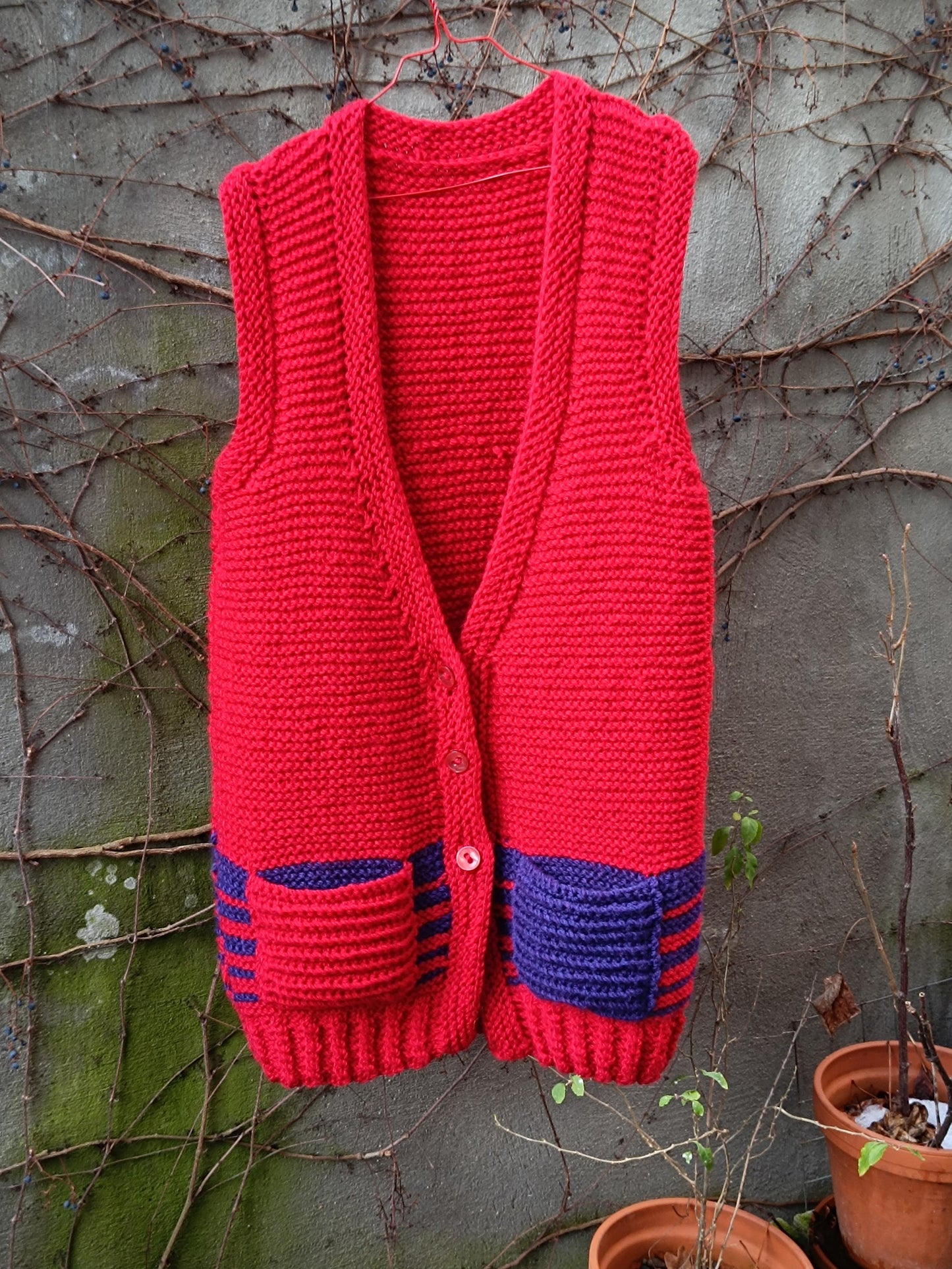 Red hand-knit sleeveless vest with purple striped pockets hanging outdoors against a vine-covered wall.