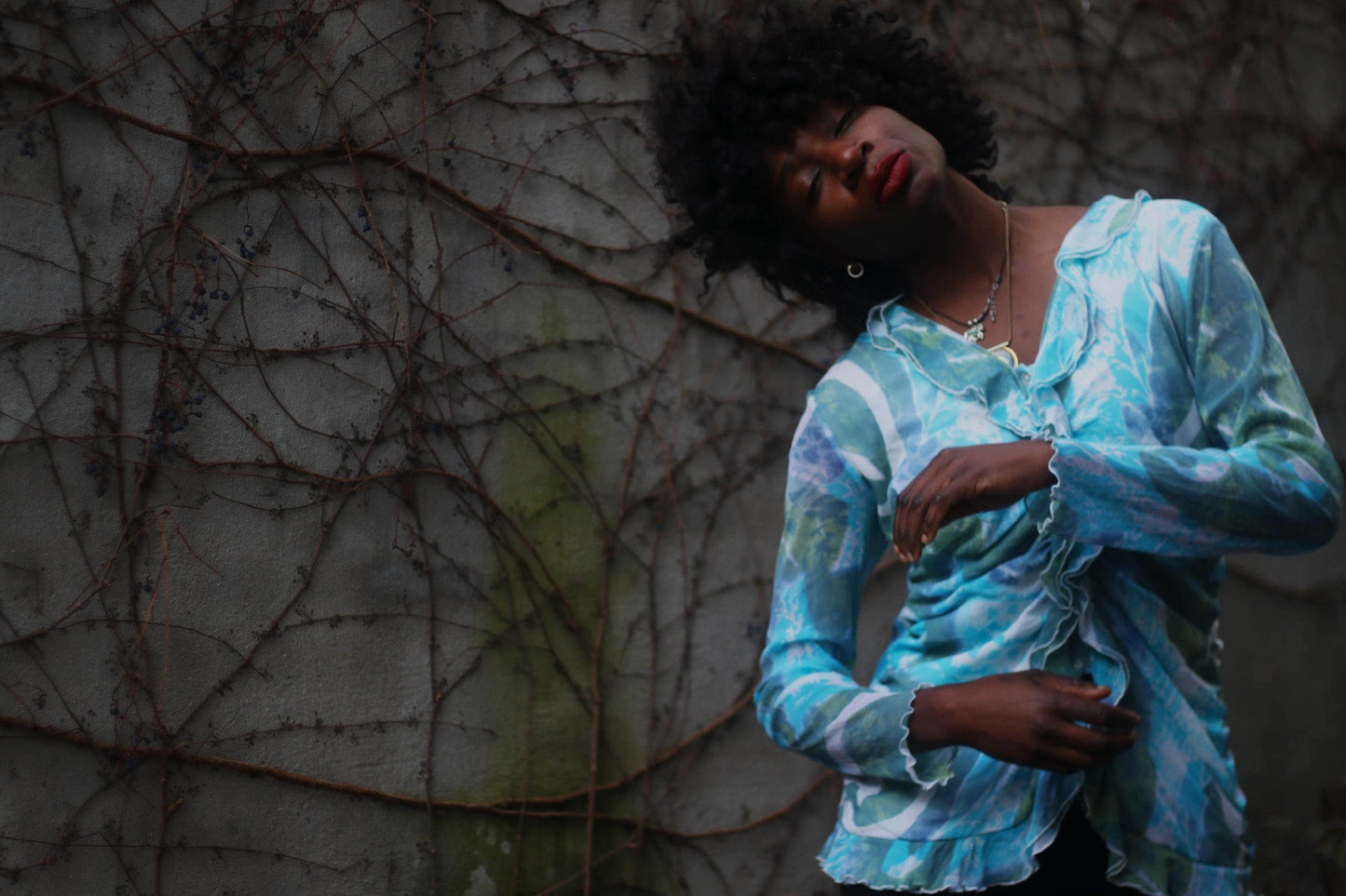 A Fashionable model of African descent in a vintage y2k blue patterned blouse posing against a textured, vine-covered wall.
