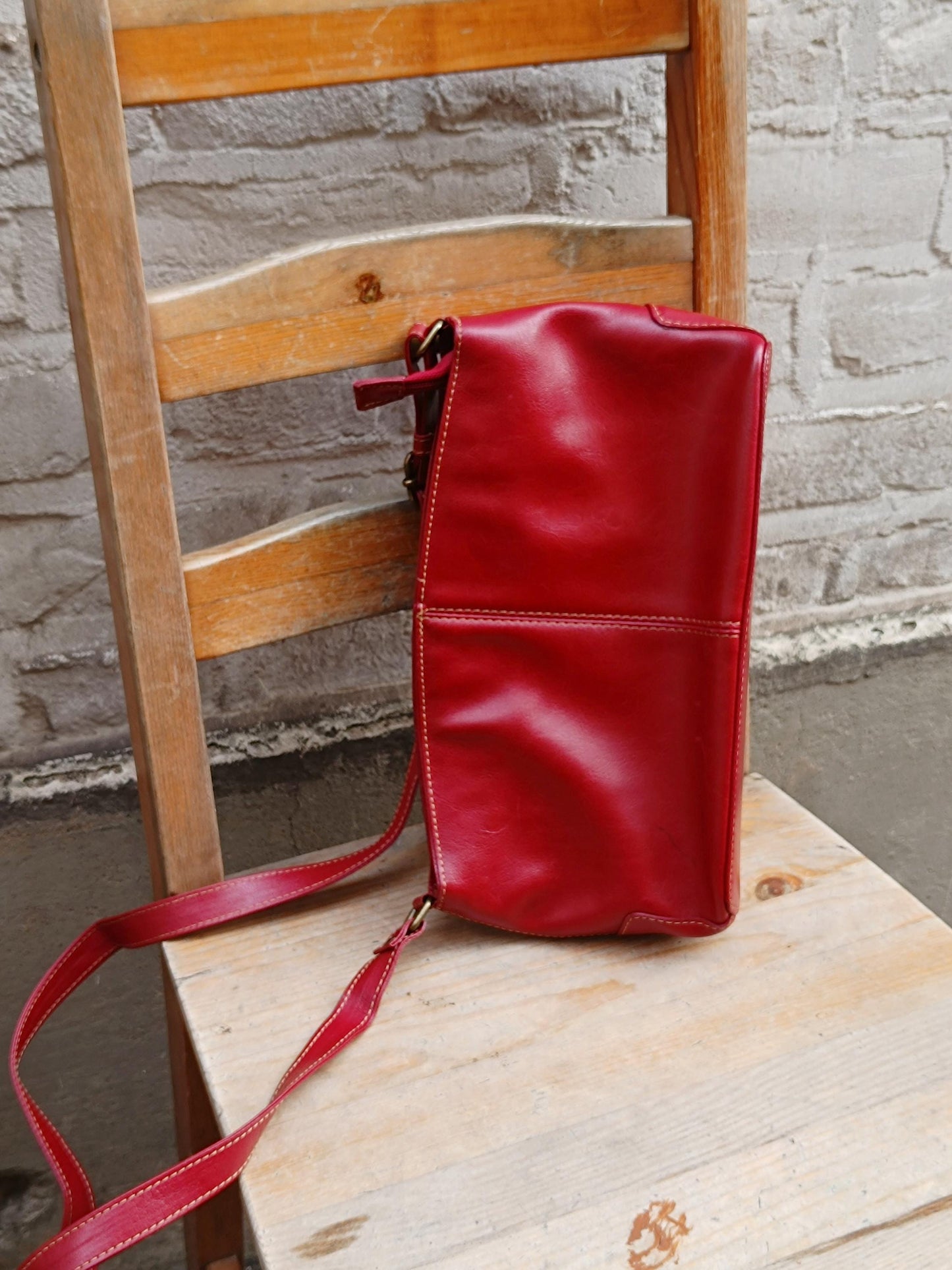 Vintage red leather shoulder bag with a single adjustable strap and silver hardware is displayed on a light wooden chair against a textured gray wall.