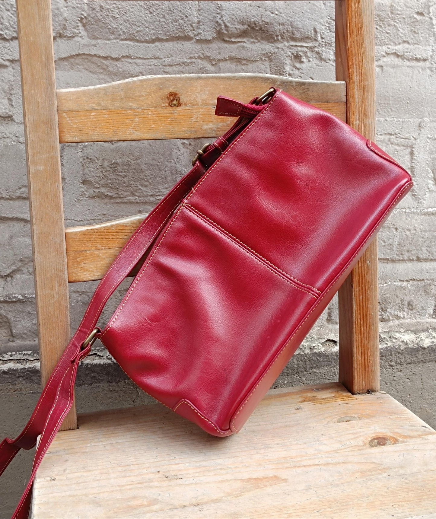 Vintage Red leather shoulder bag from the 90s is photographed leaning on a wooden chair against a white brick wall.