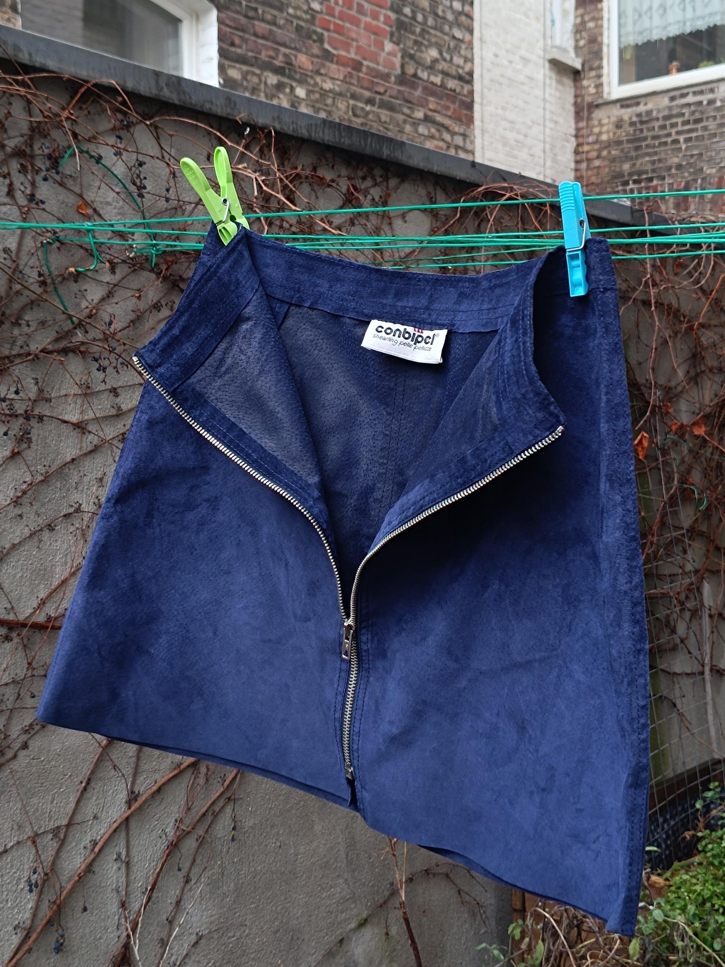 A dark blue suede skirt with a zipper down the front. The skirt is hanging on a clothesline with green clothespins. The label on the skirt reads conbipel .