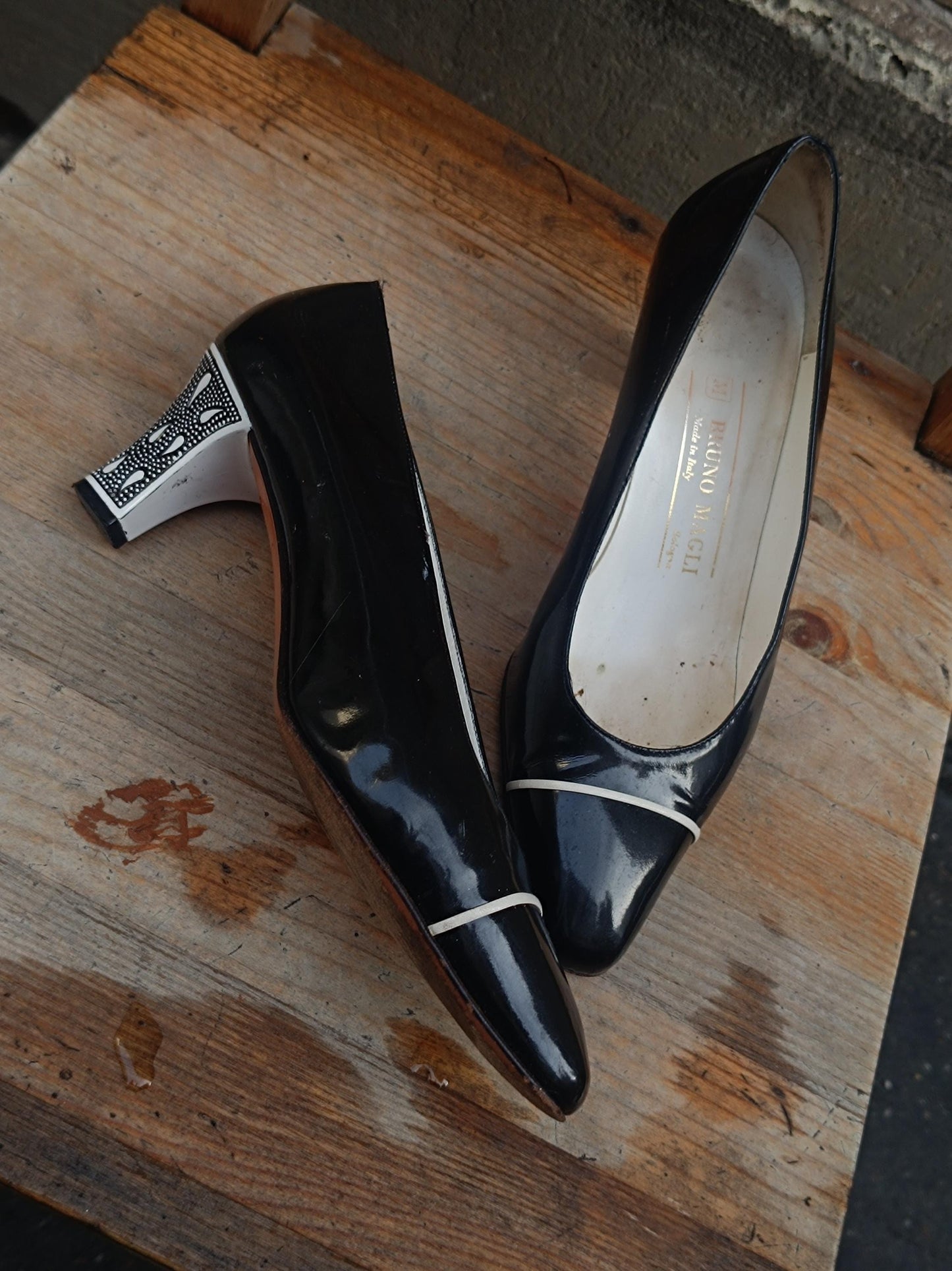 A pair of black Patent Leather Pumps by the brand Bruno Magli photographed outdoors from bird's-eye perspective on a brown rustic chair. The vintage 80s heels are statement with contrasting white heels and line in front.