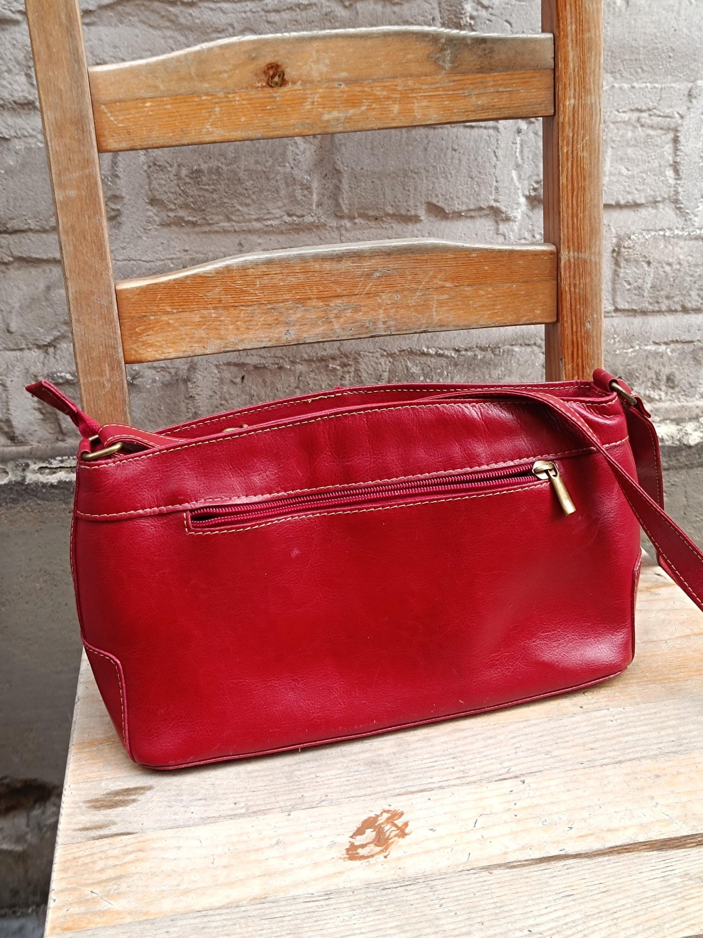 Vintage red leather shoulder bag is displayed on a light-colored wooden chair, with a textured gray wall in the background. The image provides a clear view of the bag's design, color, and condition, including its adjustable strap and silver zipper.
