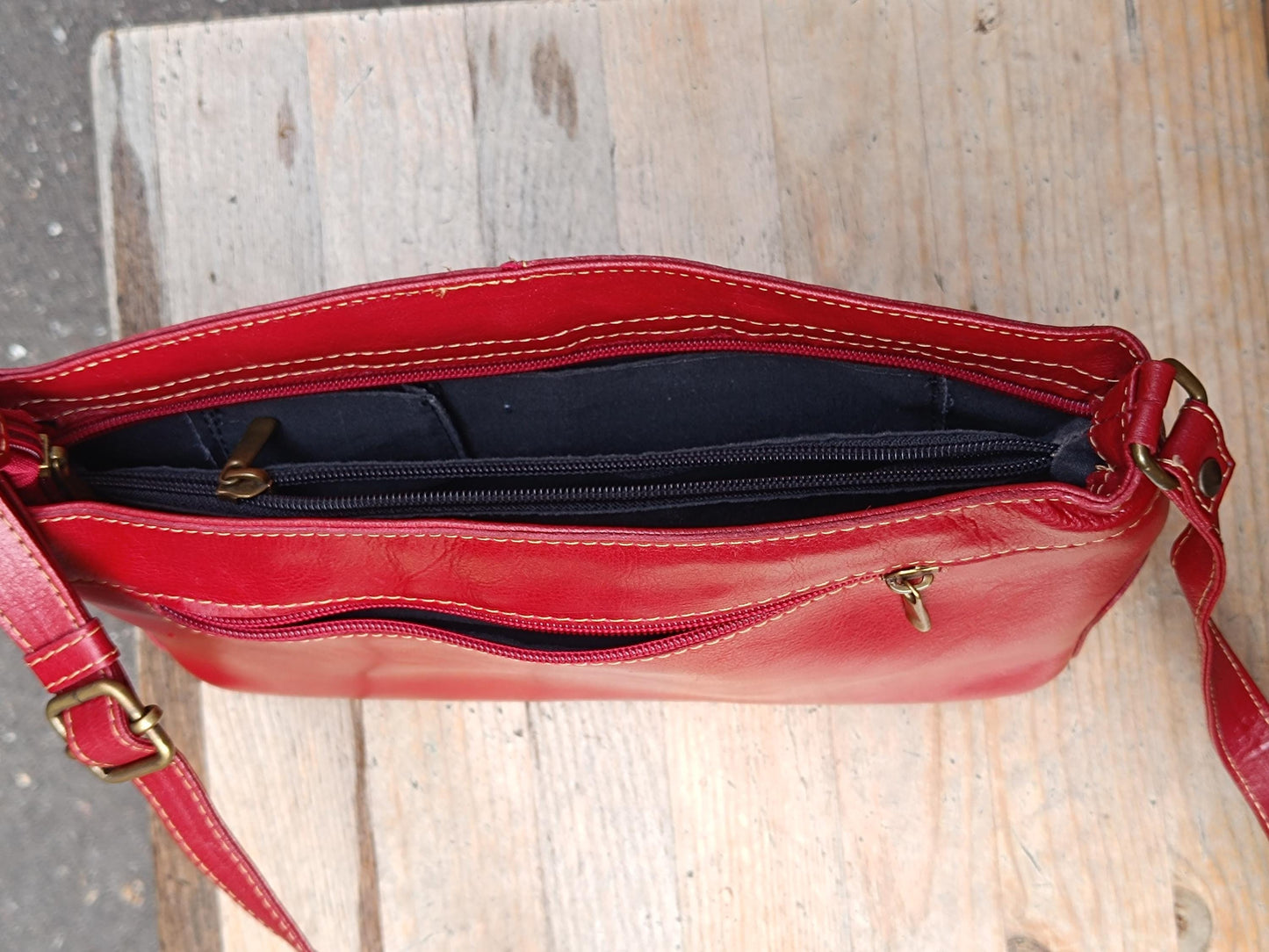 A bird's eye view of a vintage red leather shoulder bag on a light-colored wooden chair. The image provides a clear view of the inside of the bag and its multiple compartments. it also clearly shows the color, its adjustable strap, and silver zipper.