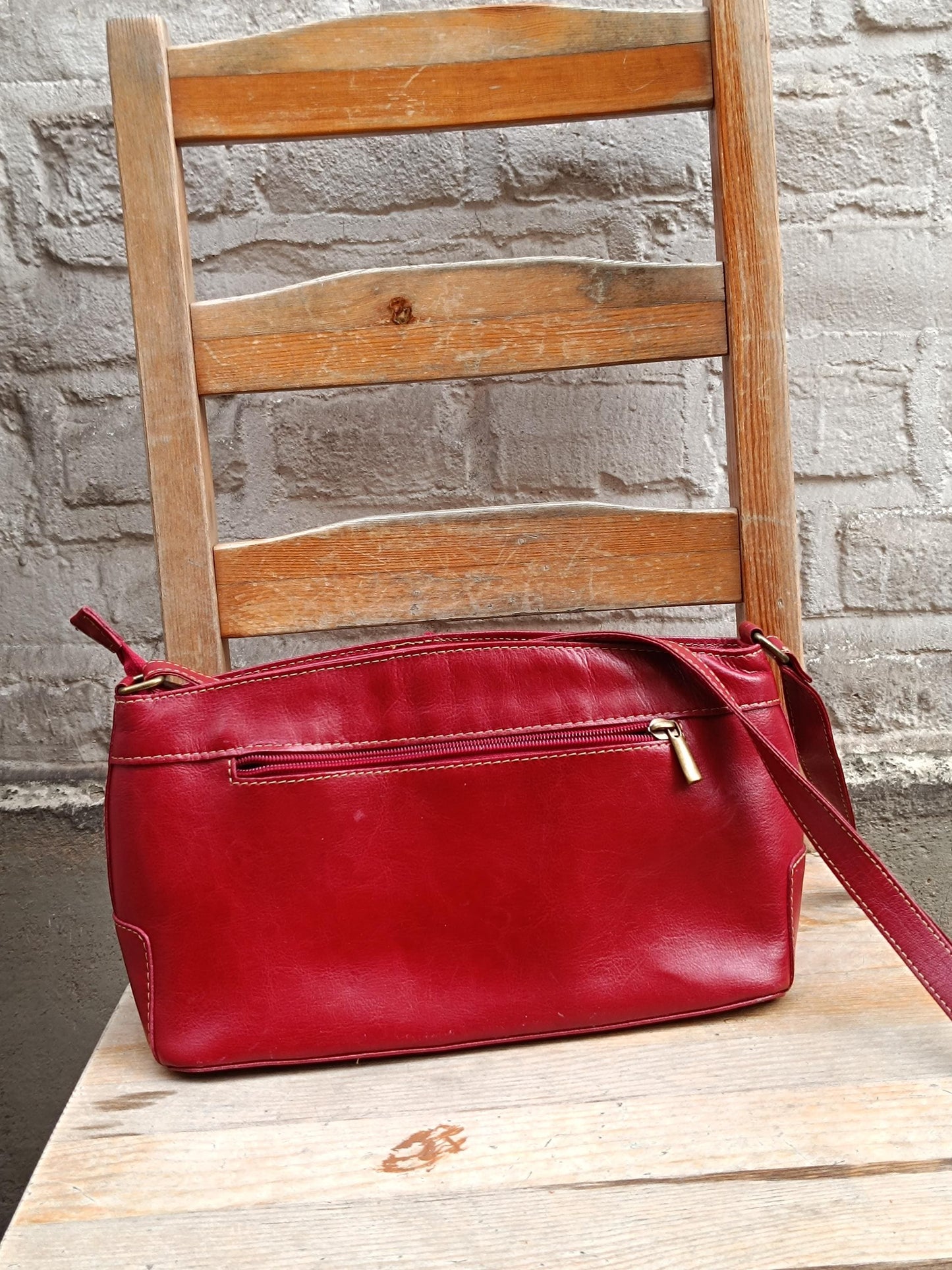 Vintage red leather shoulder bag from the 90s with a single adjustable strap and silver hardware is displayed on a light wooden chair against a textured gray wall. The bag has a visible zipper closure.