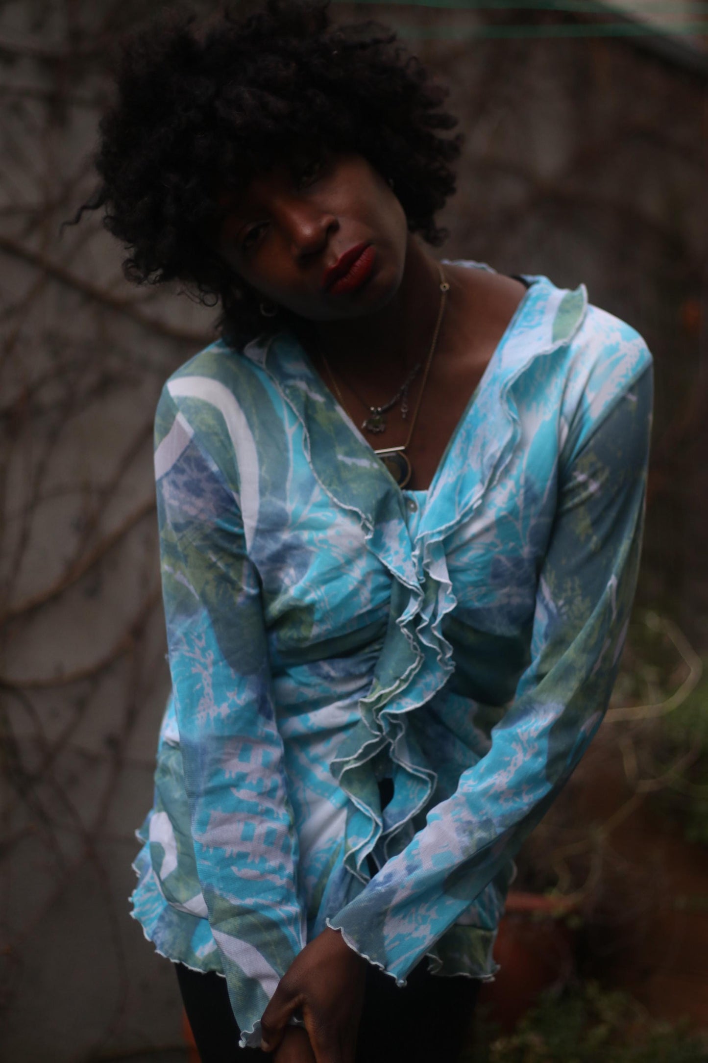 Portrait of a black female model wearing a vintage y2k patterned turquoise blouse, with a thoughtful expression, standing outdoors against a backdrop of vines.