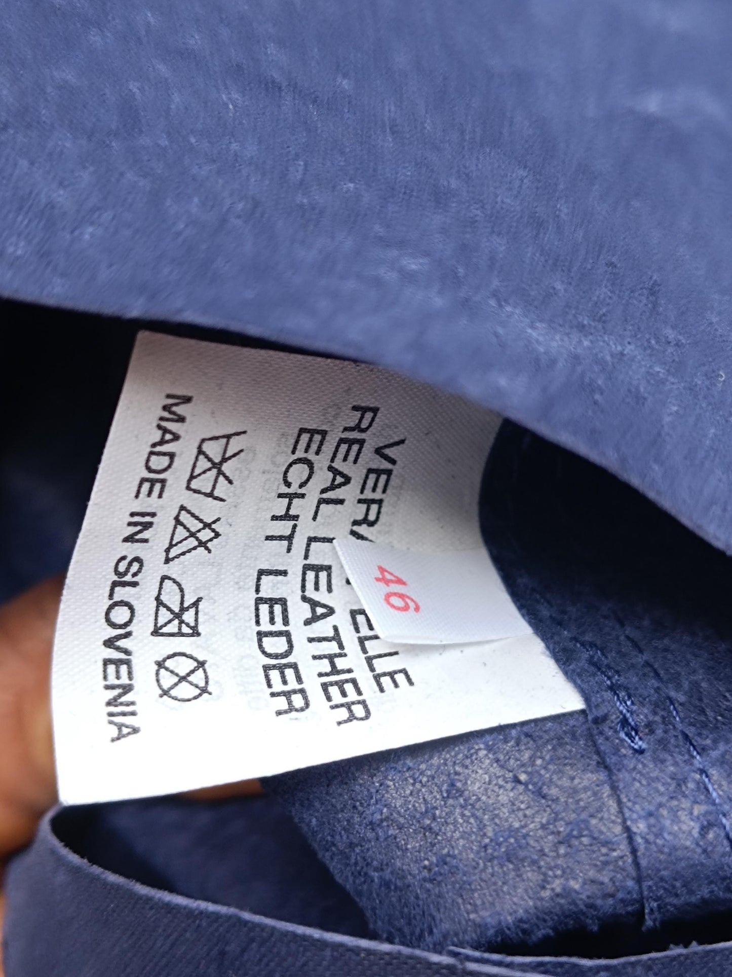 A white tag with black text that reads 'VER. 46, CELLE, REAL LEATHER, ECHT LEDER, MADE IN SLOVENIA' with a washing instructions symbol. The tag is attached to a blue suede skirt .