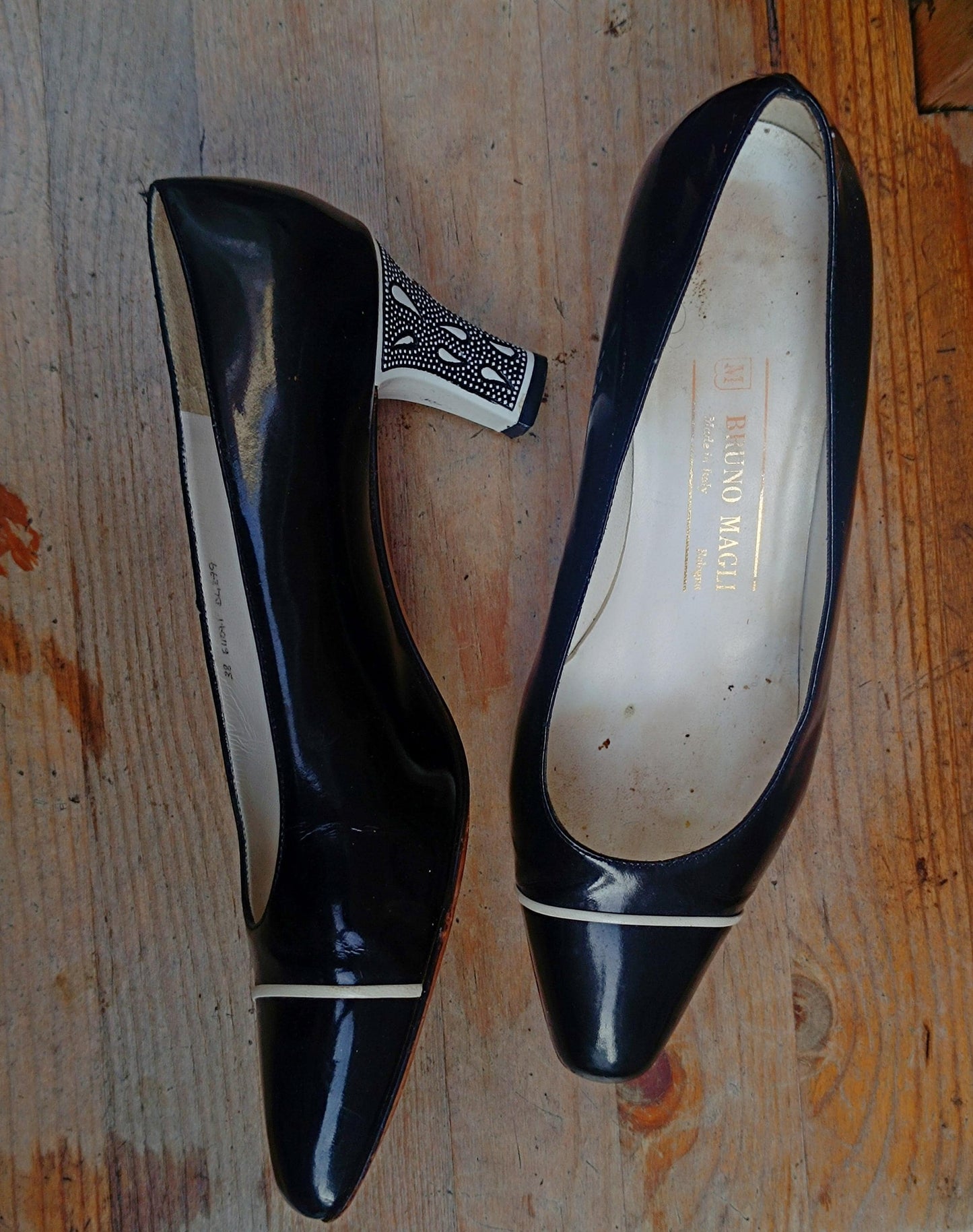 Side view of a pair of black Patent Leather Pumps by the brand Bruno Magli photographed outdoors on a brown rustic chair. The heels are statement with contrasting white heels and line in front.