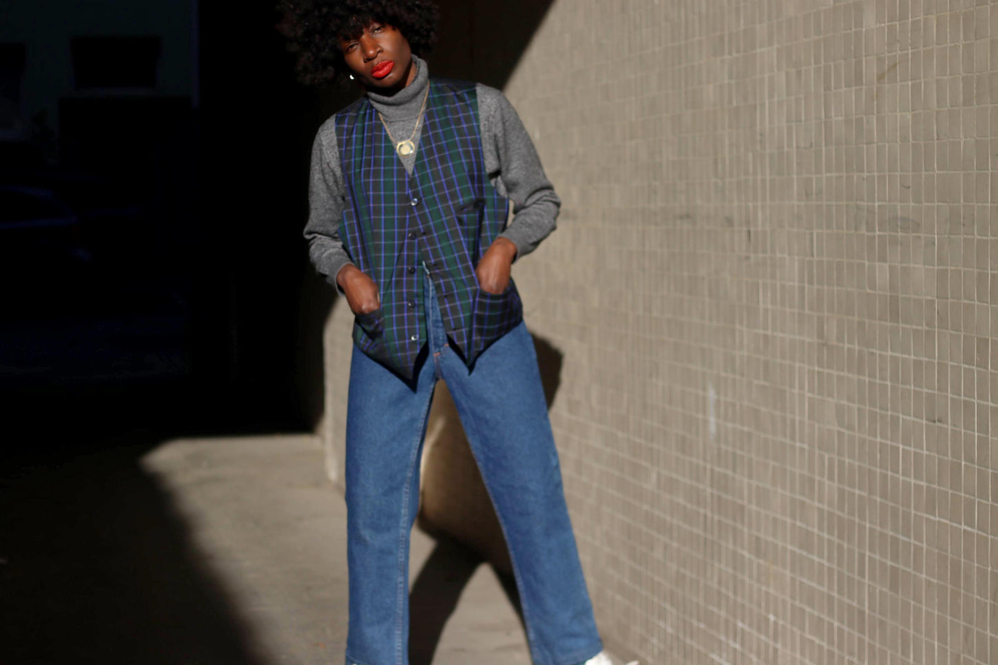 A model wearing a vintage 90s tartan plaid vest in navy and green over a gray turtleneck, styled with blue jeans, standing against a textured urban wall with dramatic sunlight.