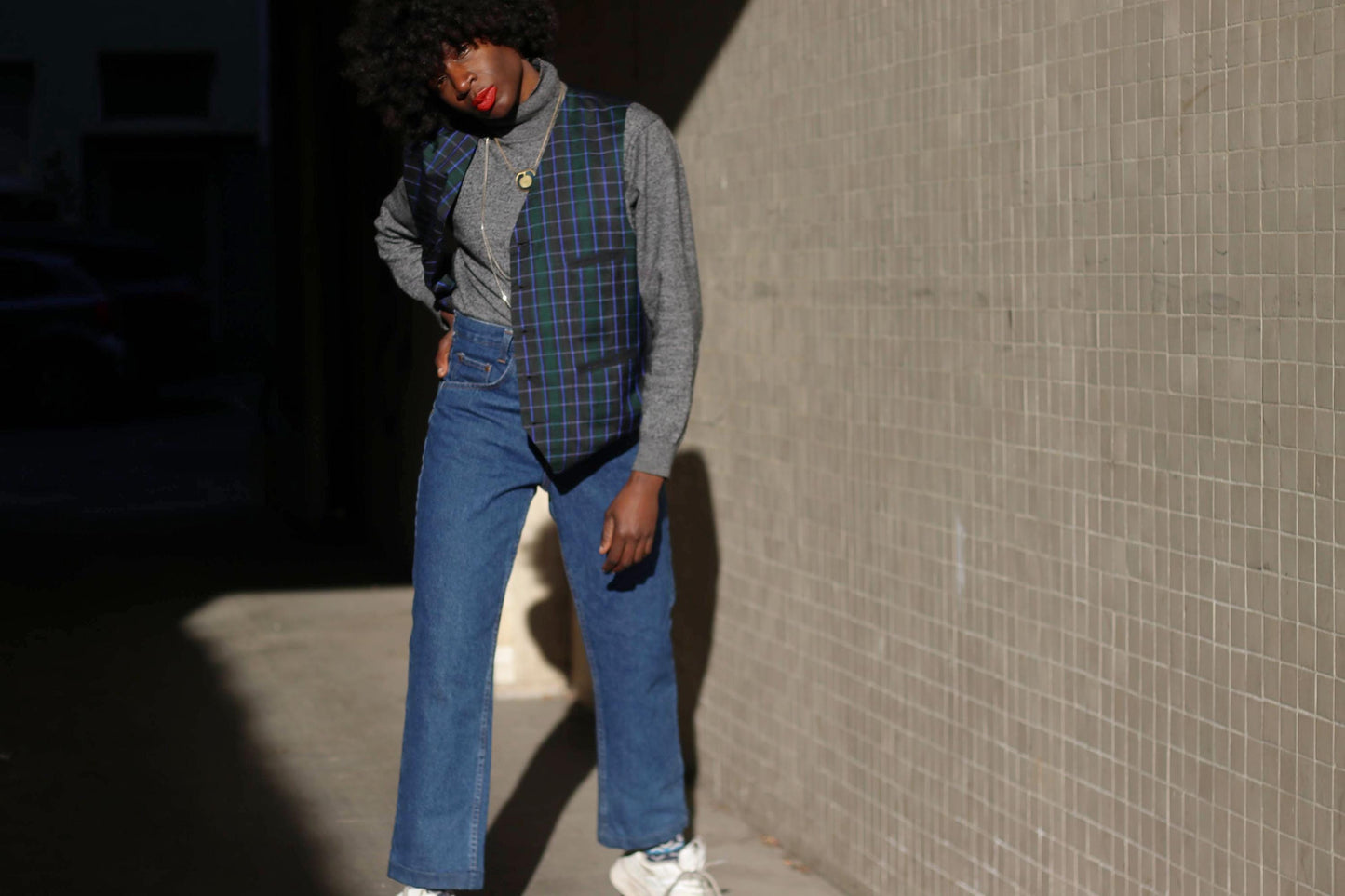 A stylish individual in a vintage plaid vest in navy and green layered over a fitted sweater, embodying a mix of preppy and grunge fashion against a modern architectural backdrop.